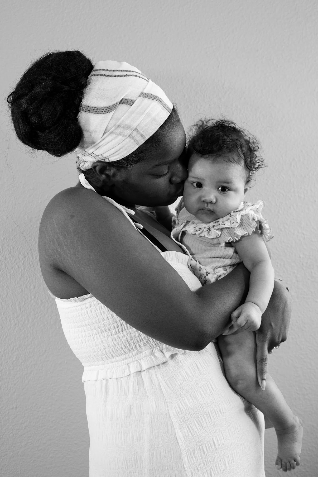 A woman holding a young child close, kissing the child's forehead, against a plain wall.