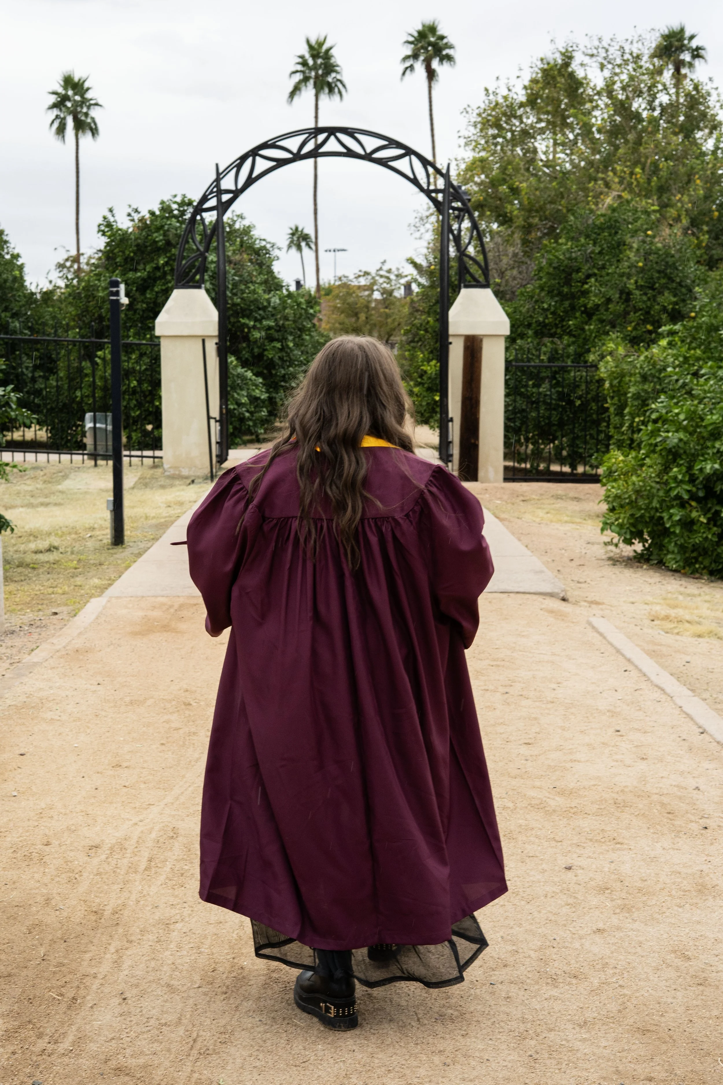 A person wearing a maroon graduation gown walking toward an archway in a park with tall palm trees.