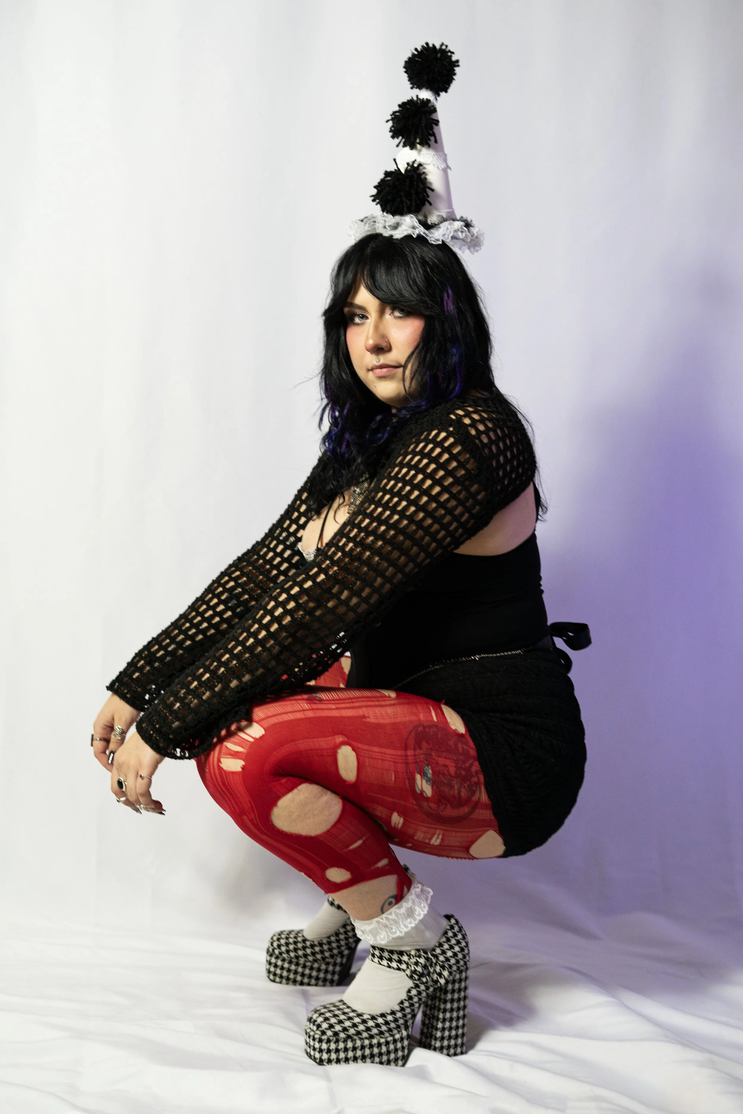 Woman squatting, wearing a black fishnet top, red tights with patterns, checkered platform shoes, and a tall, decorated party hat.