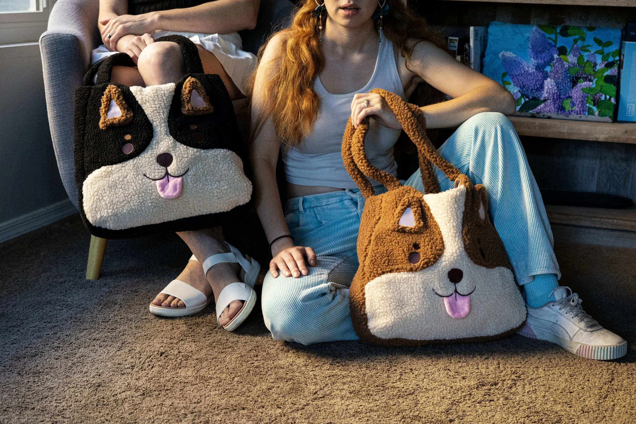 Two women sitting on the floor with plush dog-themed bags. One woman has long red hair and is wearing a white tank top and striped pants. The other woman is partially visible. The plush bags resemble the face of a dog with open mouth and tongue stick