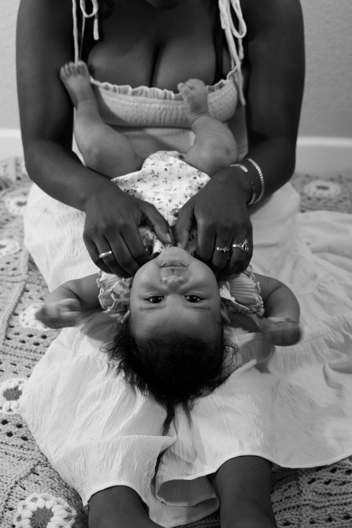 A woman sitting on a blanket holding a baby upside down, with the baby's head near her legs and feet in the air, on a crocheted rug.
