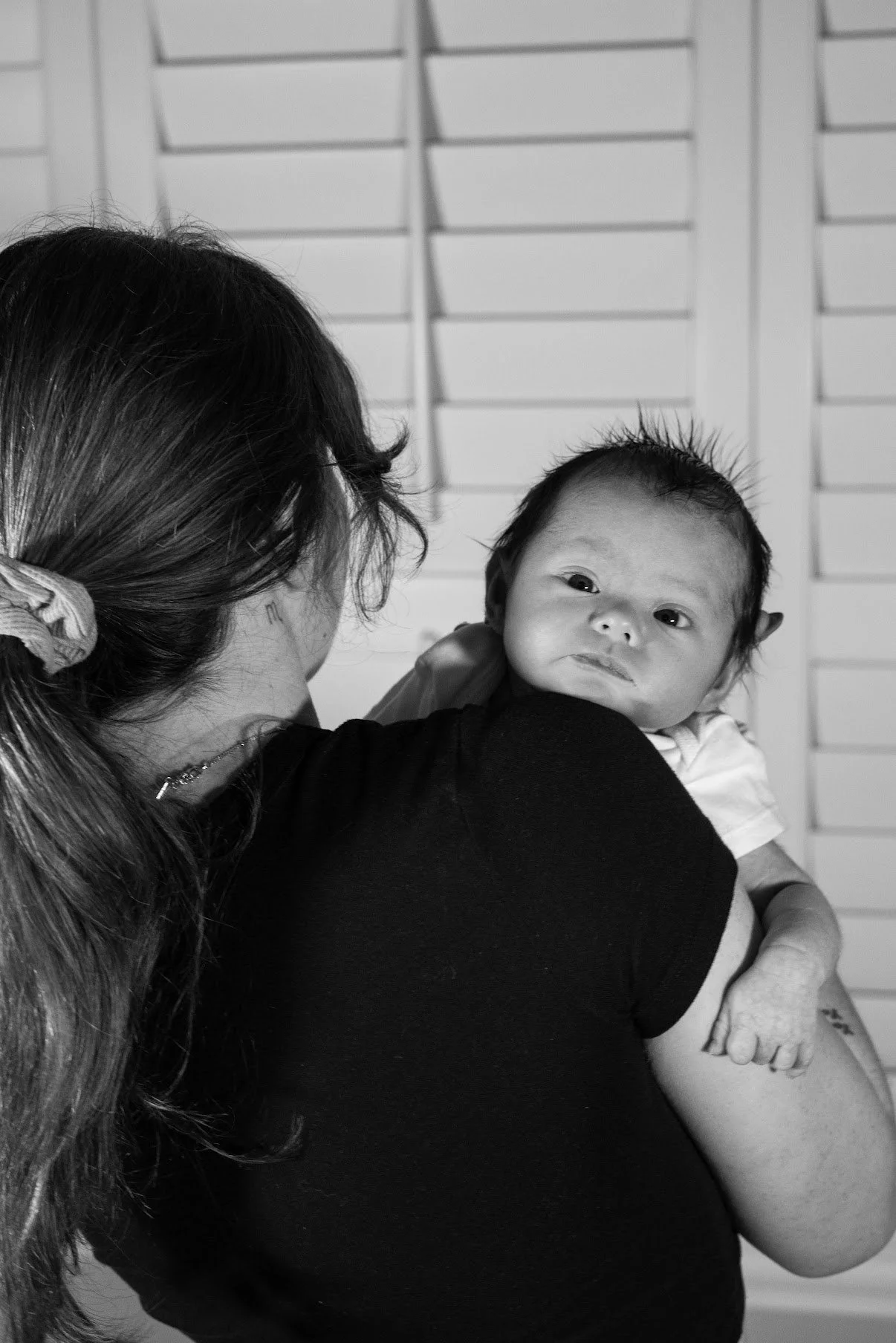 A woman holding a baby, with her back to the camera. The baby is looking over her shoulder towards the camera.