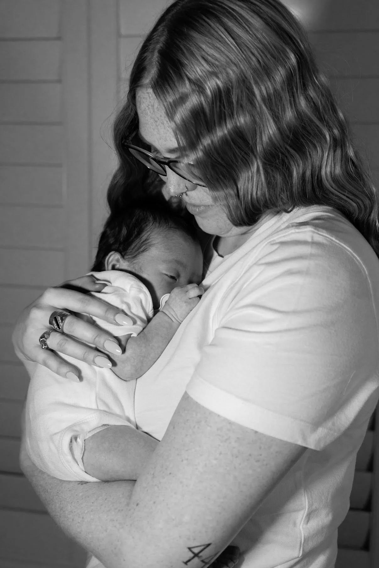 A woman with glasses holding a sleeping baby close to her chest indoors.