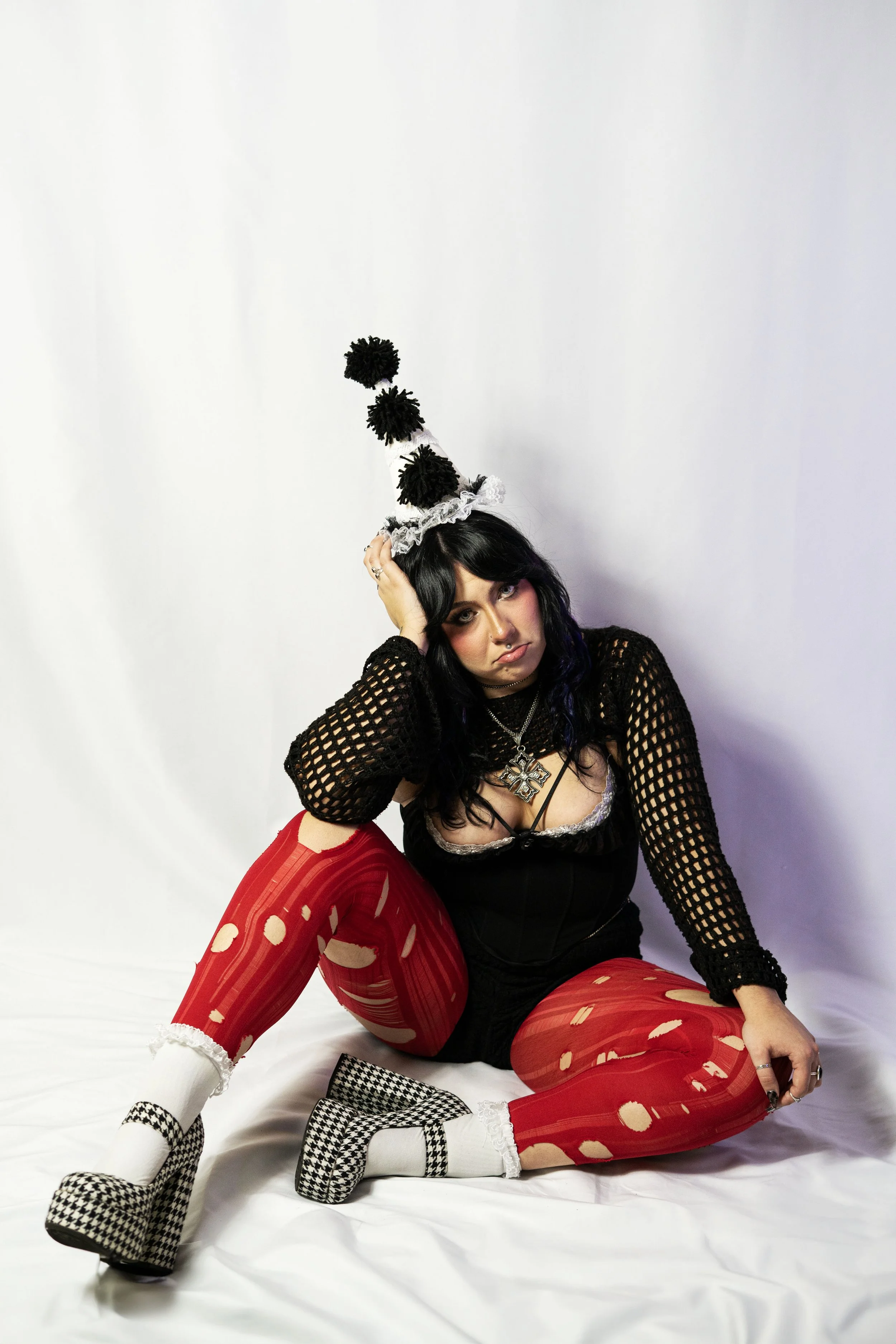A woman sitting on the floor in front of a plain white background. She wears a black netted long-sleeve top, a black corset, red patterned tights, white socks, and houndstooth platform shoes. She has dark, wavy hair, and accessorizes with a large cro