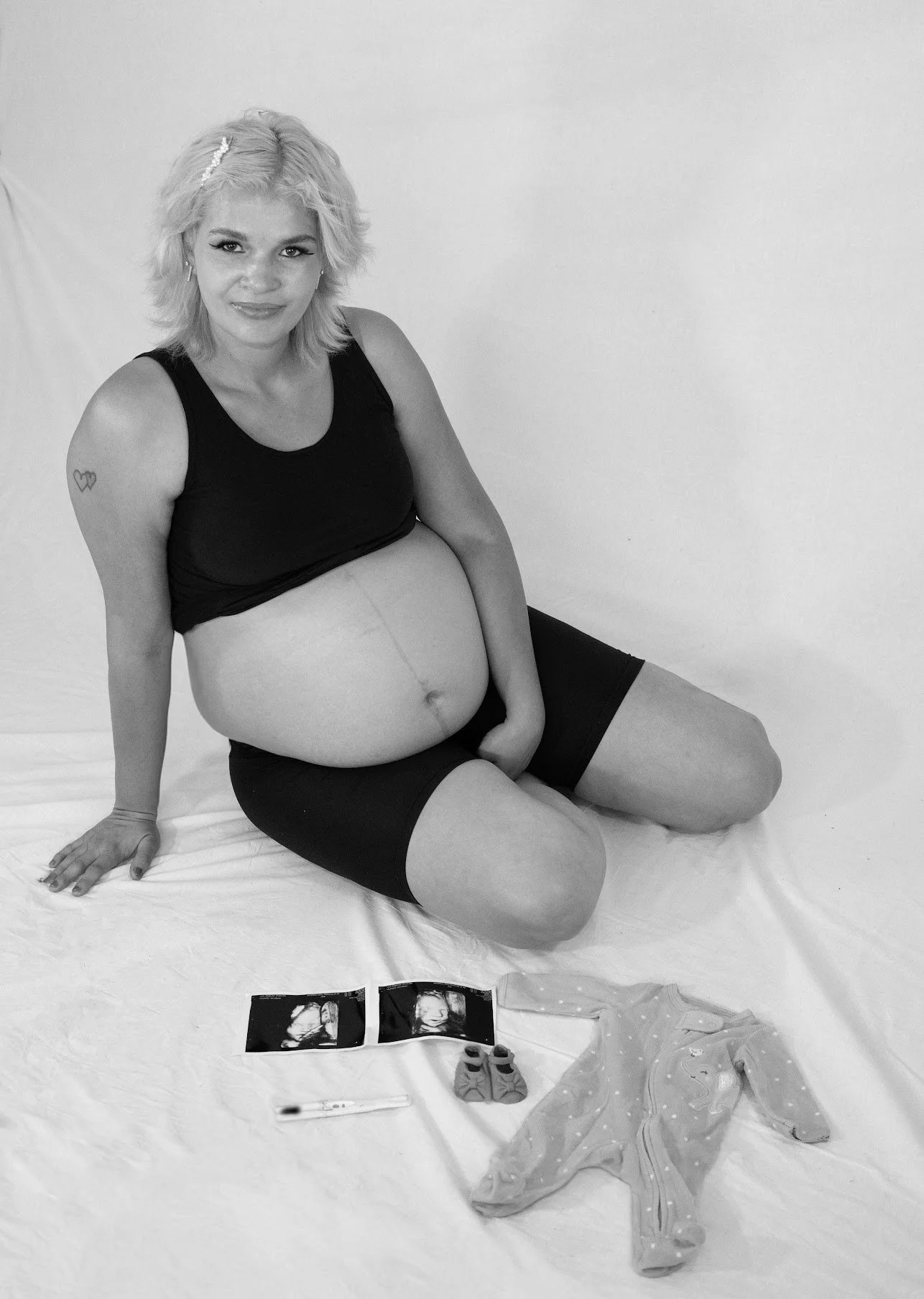 A pregnant woman sitting on a white surface, with ultrasound pictures, baby sock, a onesie, and a thermometer in front of her.