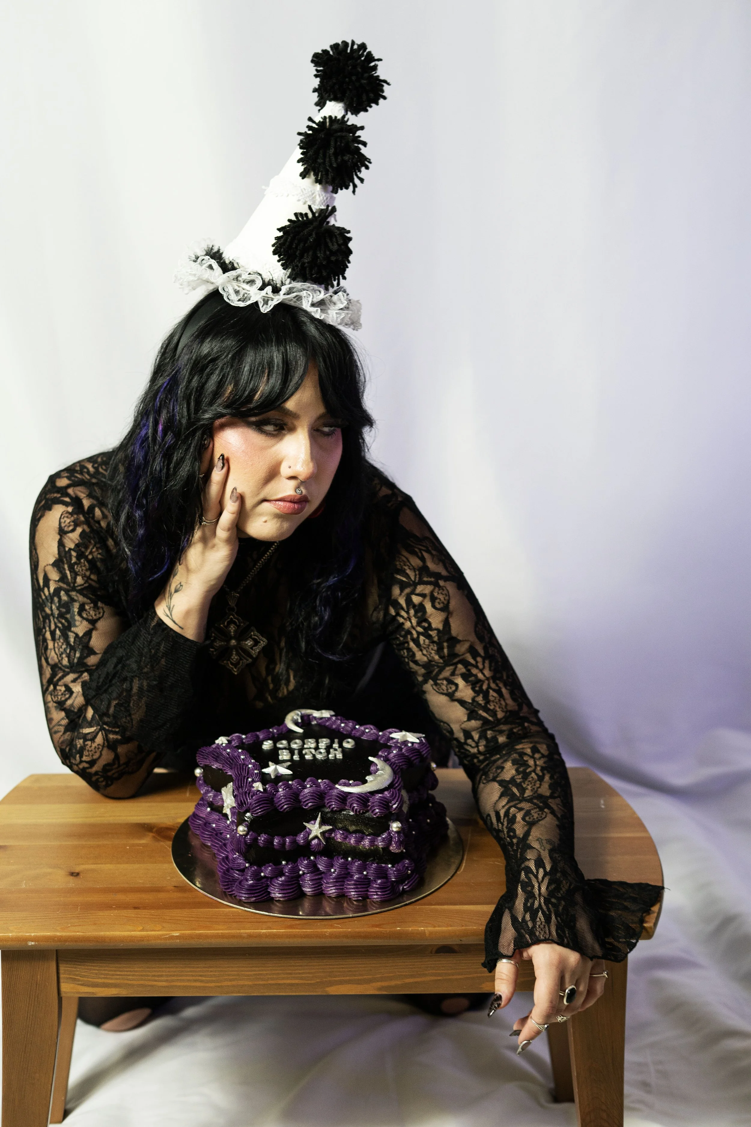 A woman with black hair and tattoos, wearing a black lace outfit and a pointed birthday hat with black pom-poms, sits at a wooden table with a purple birthday cake decorated with silver stars and crescent shapes.