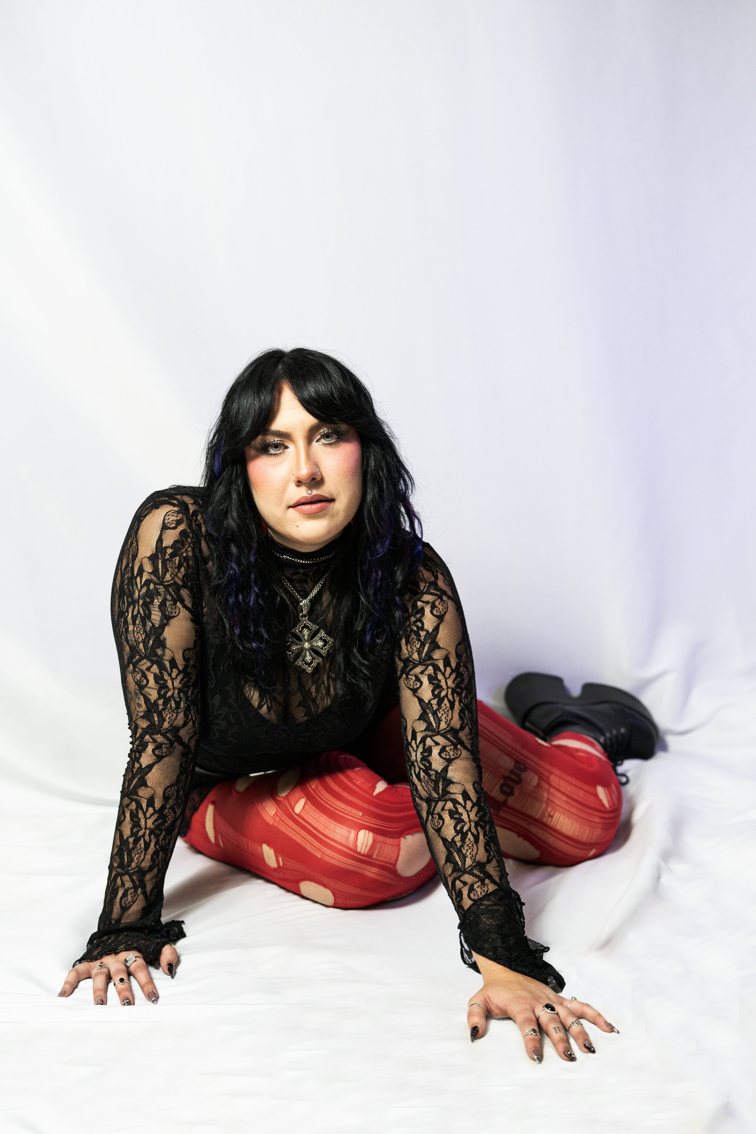 A woman with black hair and makeup, wearing a black lace top, layered necklaces, and red patterned tights, is posing on her hands and knees against a white background.