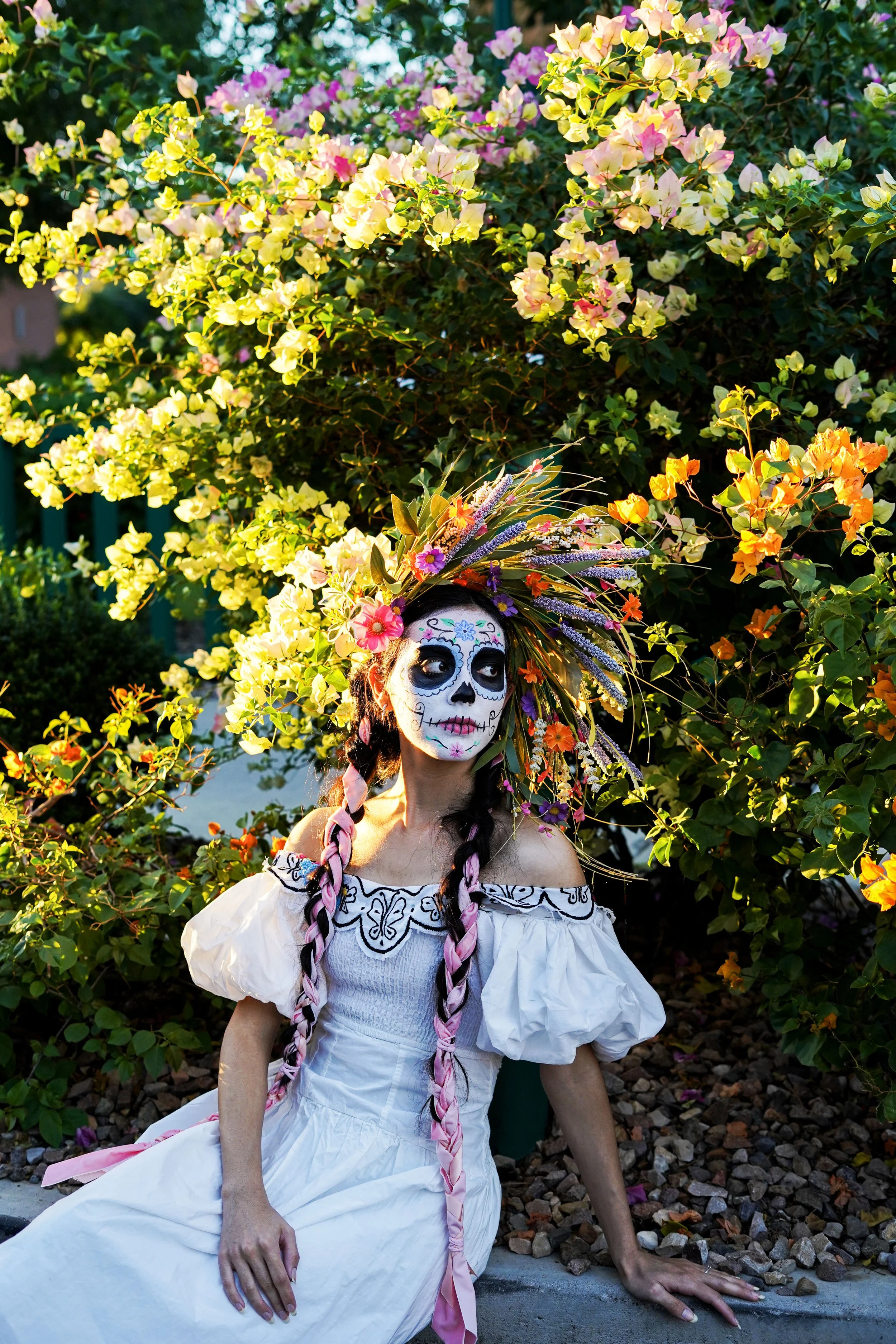 Woman in traditional Mexican dress with sugar skull face paint, wearing a colorful floral headdress, sitting outdoors near a flowering bush.