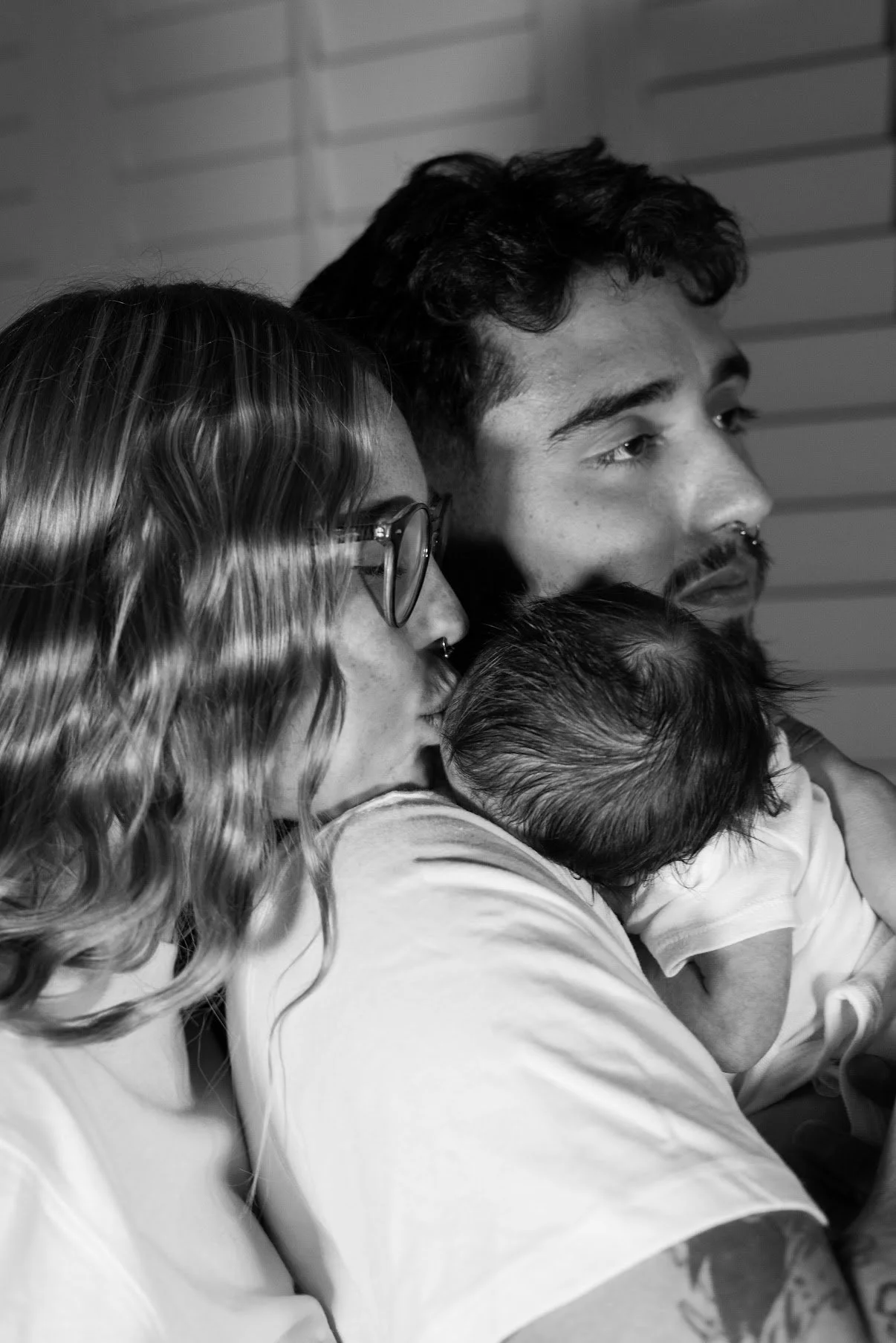 A black and white photo of a family showing a woman with glasses and wavy hair, a man with a beard and curly hair, and a baby with wet hair all close together, possibly in a hospital or care setting.