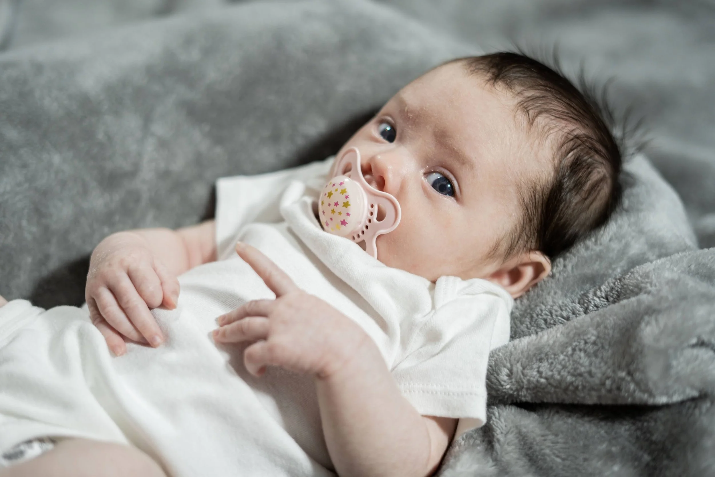 Close-up of a baby with blue eyes lying on a soft gray blanket, wearing a white outfit, with a pink pacifier with star patterns in their mouth.