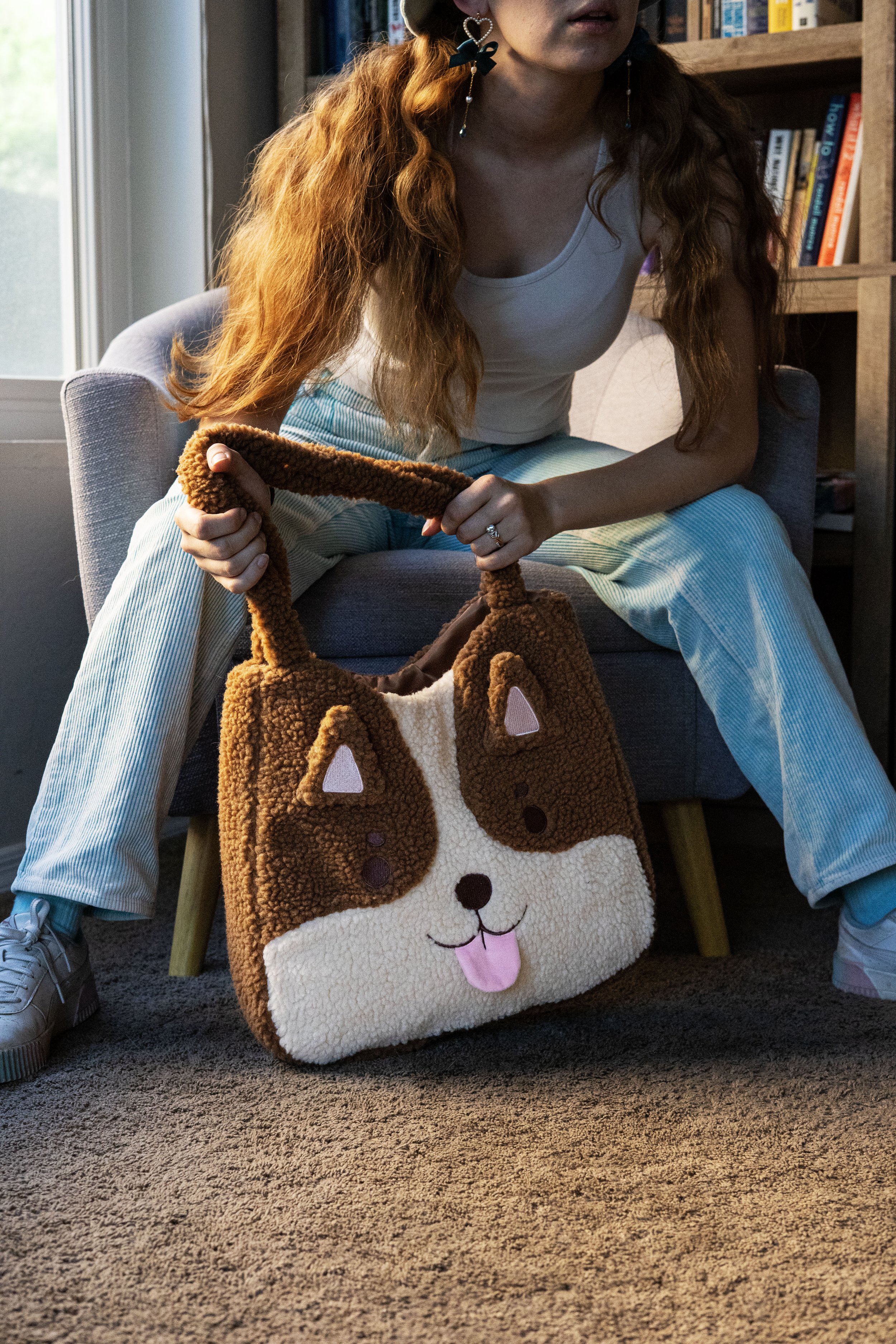 A woman with red curly hair wearing earrings, a white tank top, and light-colored striped pants, sitting on a gray armchair, holding a brown and white dog-shaped plush bag with a cartoon face and tongue sticking out, in a room with bookshelves and a window nearby.