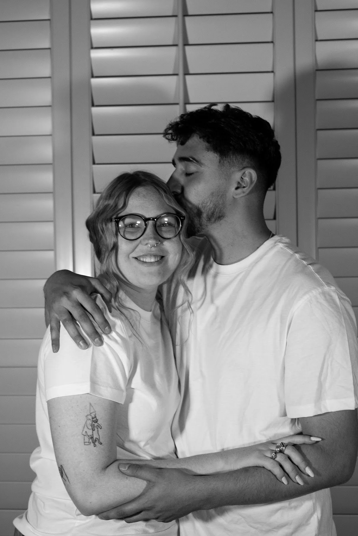 A woman with glasses and tattoos is embracing a man who is kissing her on the forehead. They are smiling and standing in front of a shuttered door.