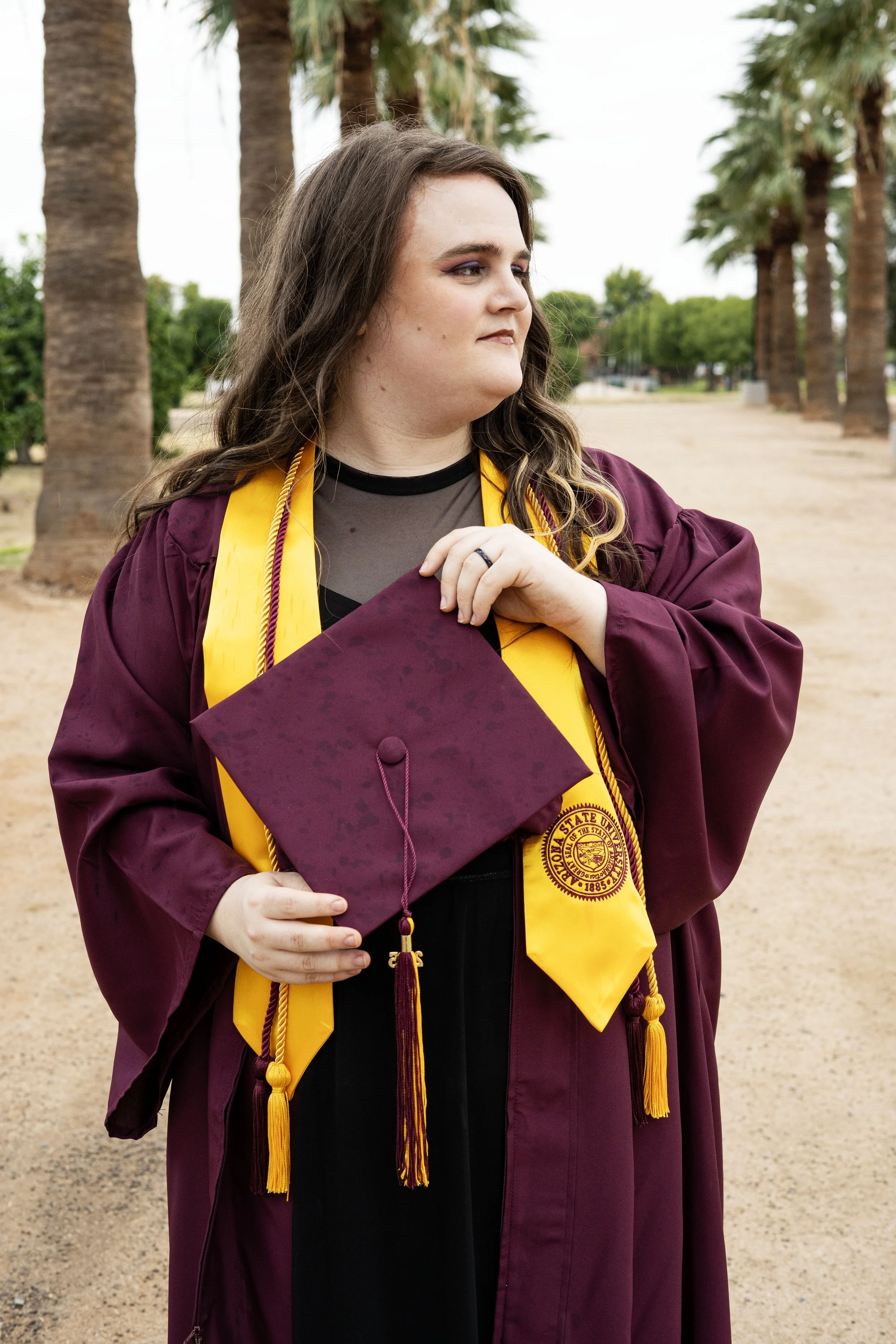 A woman in a maroon graduation gown and yellow stole holding a maroon cap, standing outdoors on a pathway lined with palm trees.