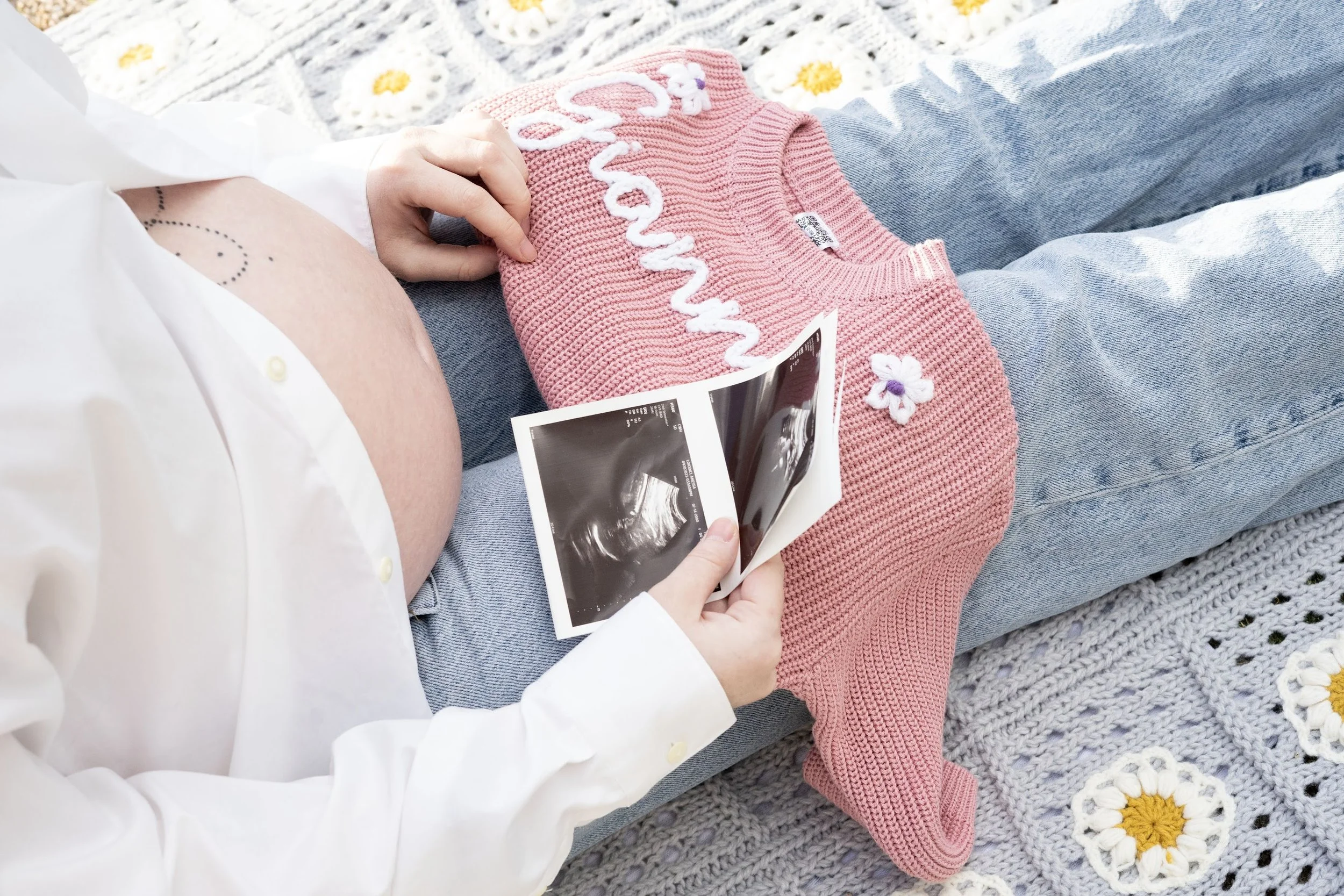Pregnant woman lying on her side holding ultrasound photos, with a pink knitted sweater and daisies on a knitted blanket nearby.