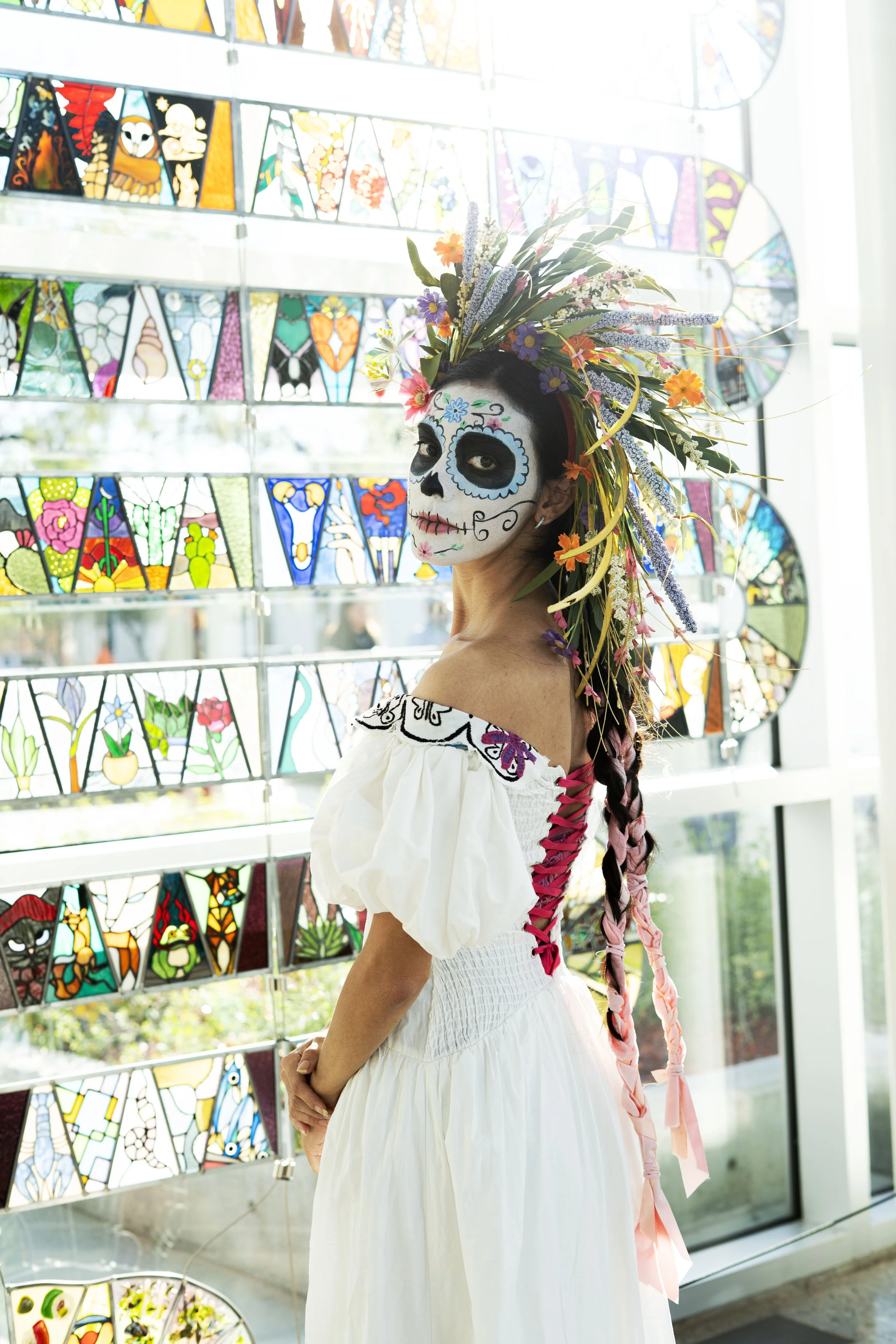 Woman dressed as a calavera for Día de los Muertos, wearing a white dress with puffed sleeves and a decorated face with skull makeup. She has long braided hair and a large floral headpiece with colorful flowers and greenery. Behind her are stained gl