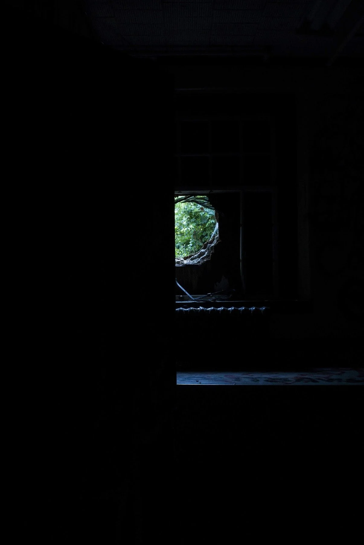 The photo is completely dark, we are looking into a hole in the wall. It is craved out of brick, we see faint light trickling into the room, the hole in the wall is bright and you can see a garden on the other side.