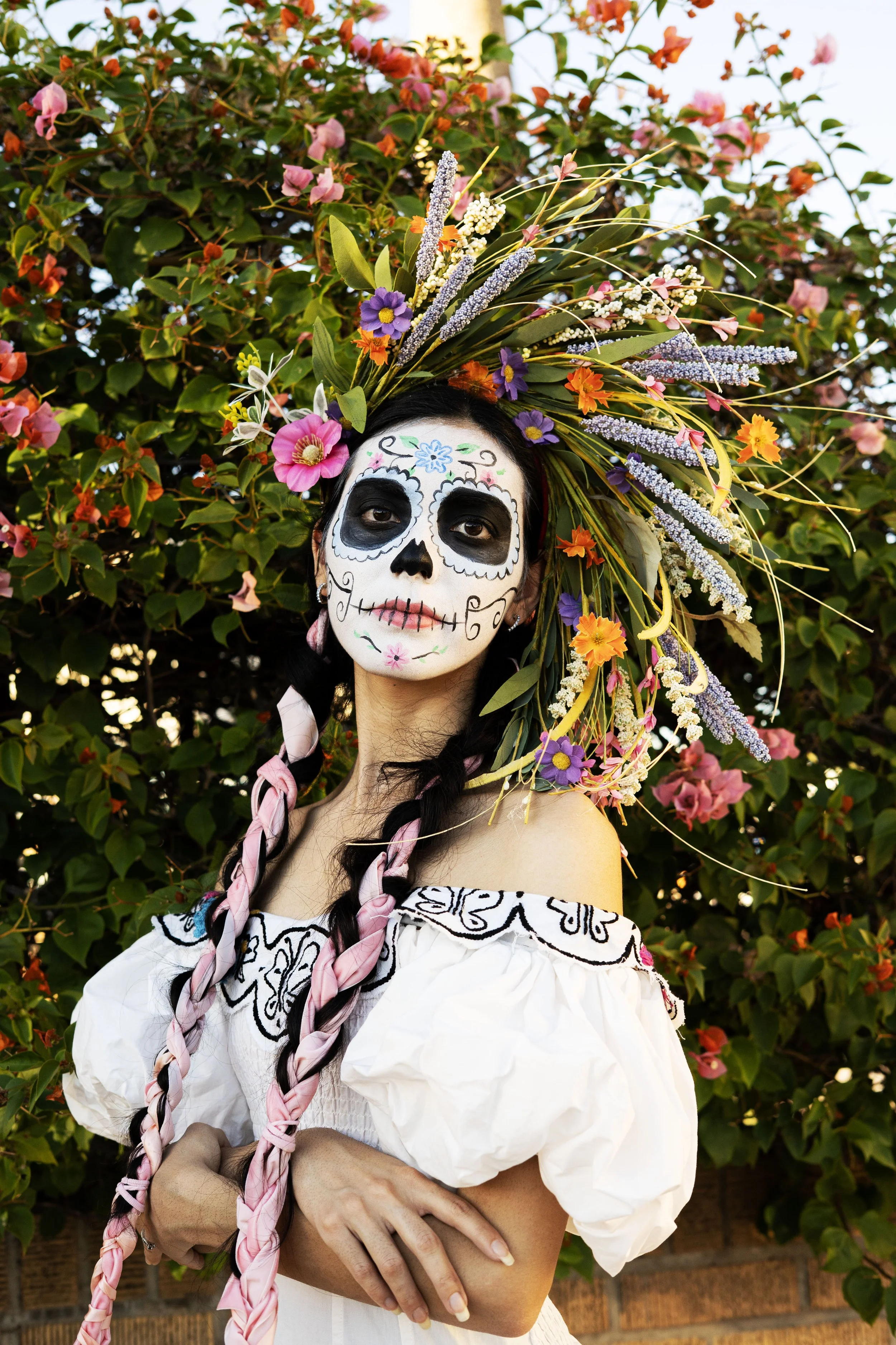 A woman dressed in traditional Mexican attire with sugar skull face paint, wearing a colorful floral headdress, standing outdoors near flowering bushes.