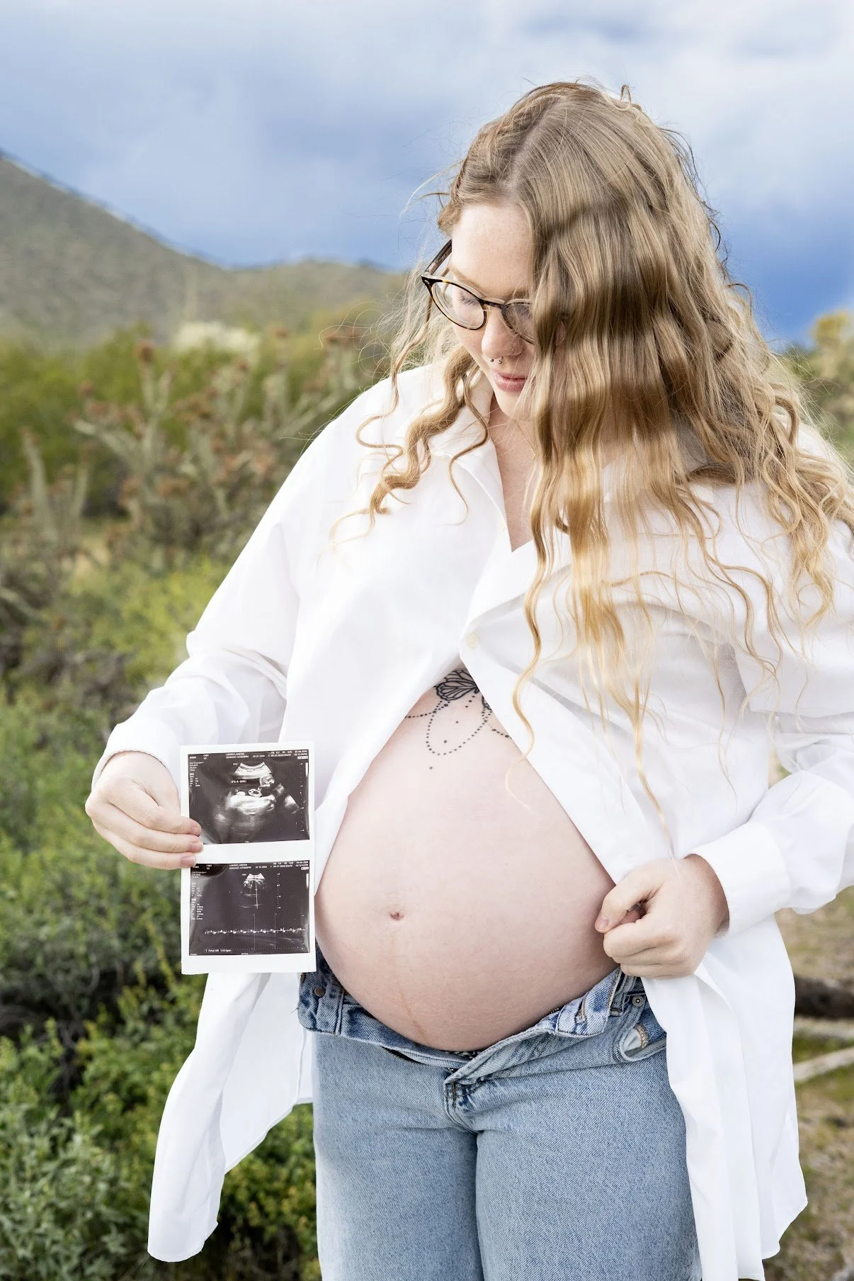A pregnant woman with long curly red hair and glasses holding an ultrasound picture outdoors, smiling and looking down at her belly.