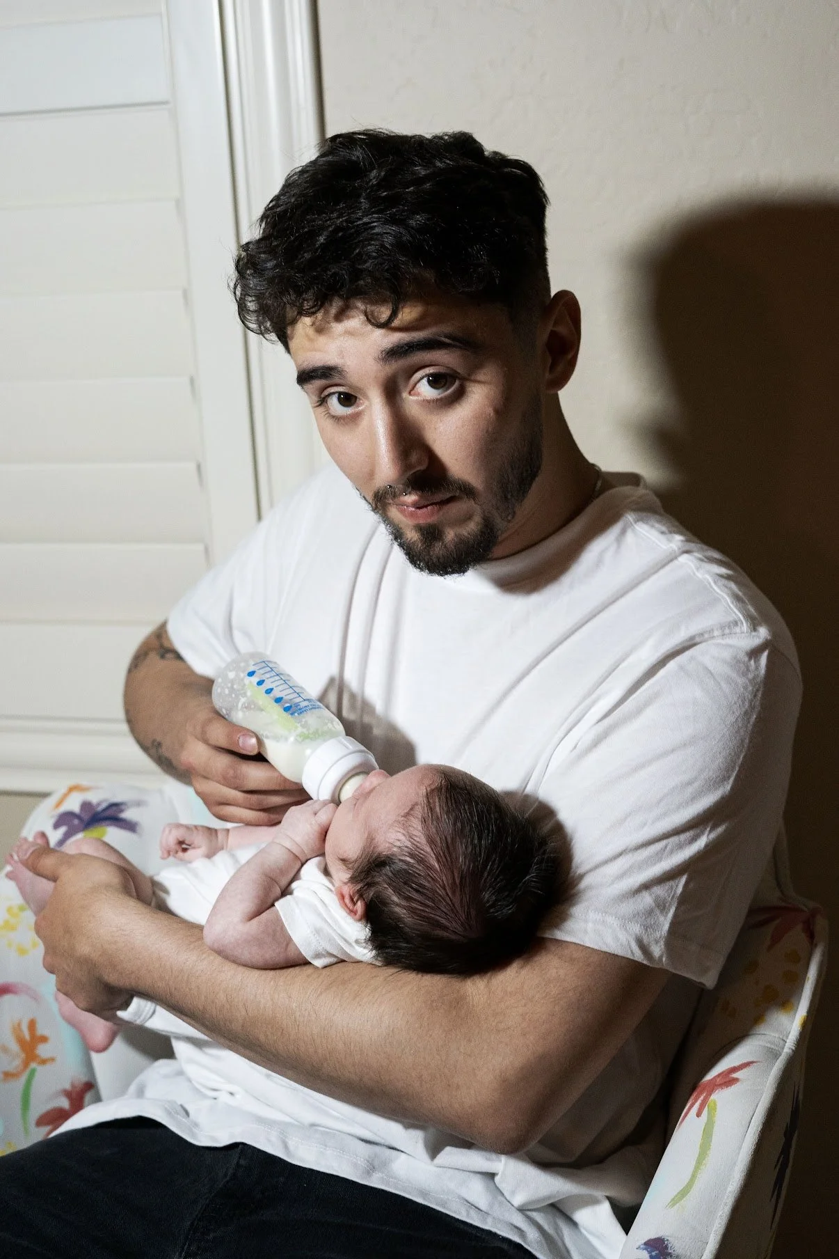 A man with dark hair and a beard feeding a newborn baby with a bottle while sitting on a chair.