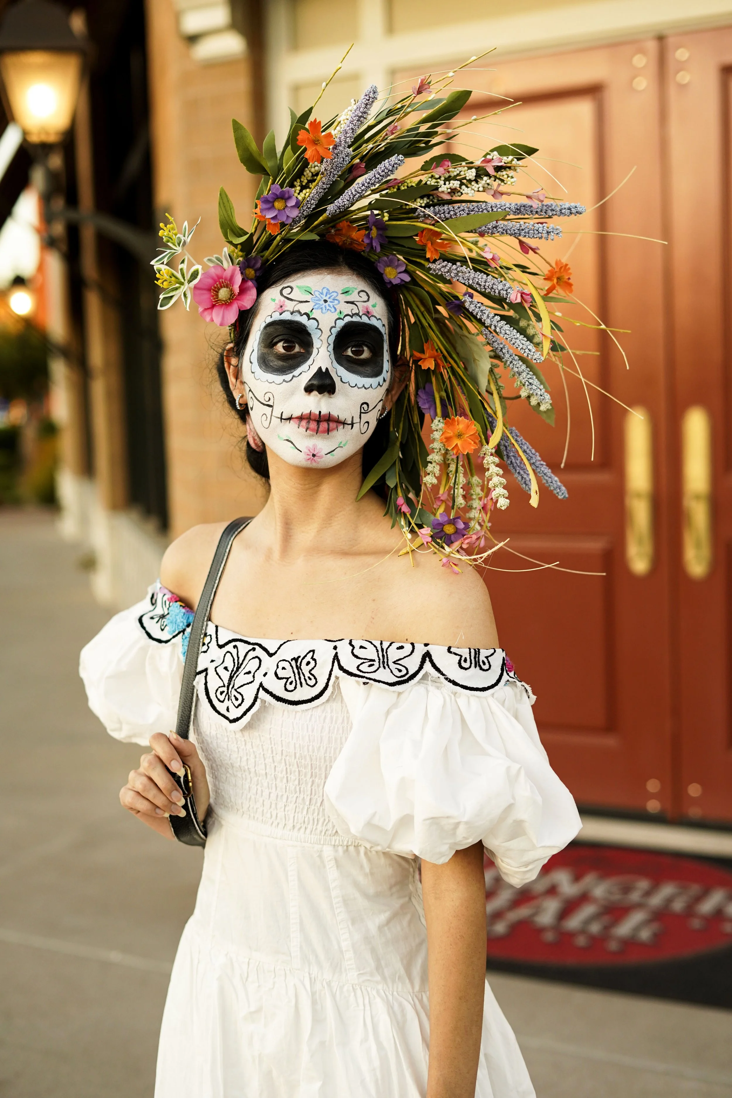 Woman dressed in a white dress with off-the-shoulder puffed sleeves and black embroidery, wearing a colorful floral headpiece and sugar skull makeup for Día de los Muertos.