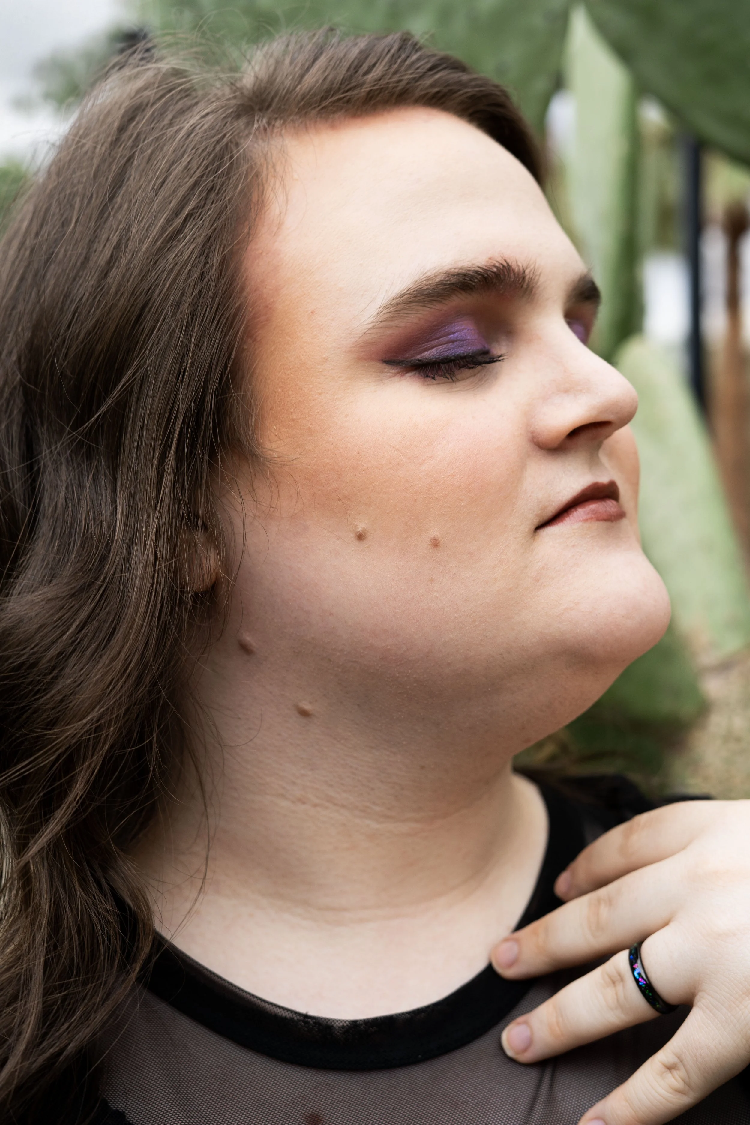 A woman with long brown hair and purple eyeshadow, her eyes closed, touching her neckline with her right hand, wearing a black ring and a black sheer top, outdoors with green plants in the background.