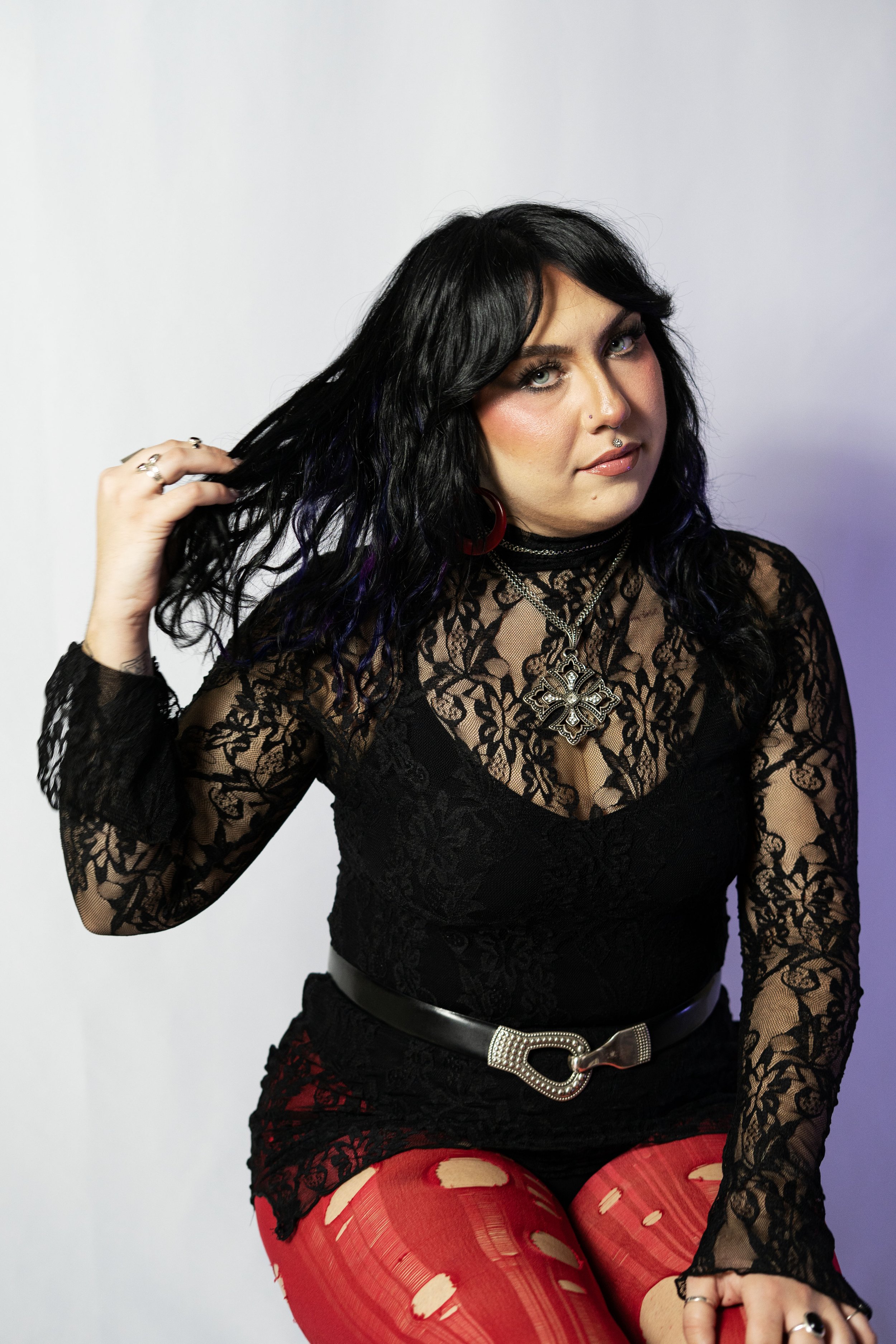 A woman with long black hair and blue eyes, wearing a sheer black lace top, red ripped tights, and red hoop earrings, sitting against a plain white background.