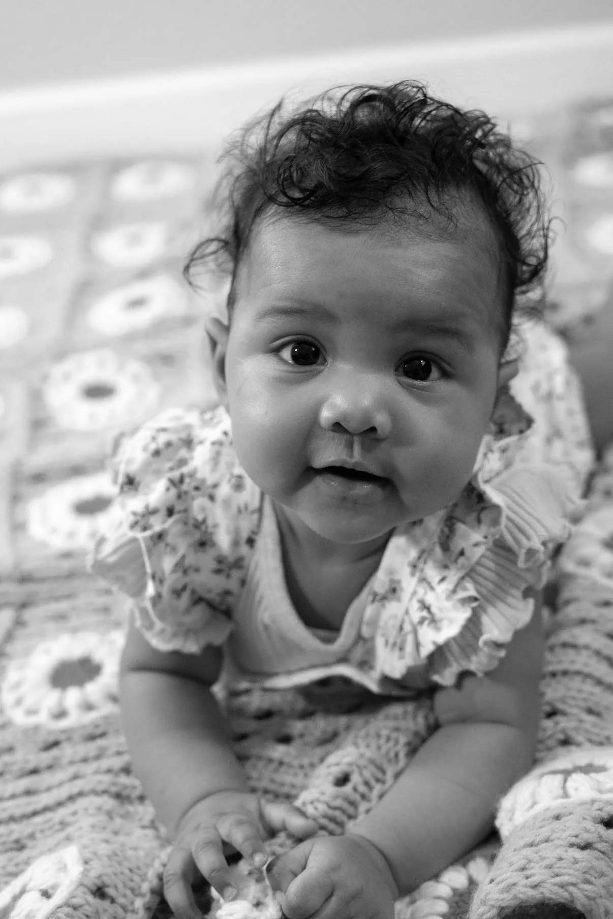 A baby with curly hair looking directly at the camera, lying on a patterned blanket.