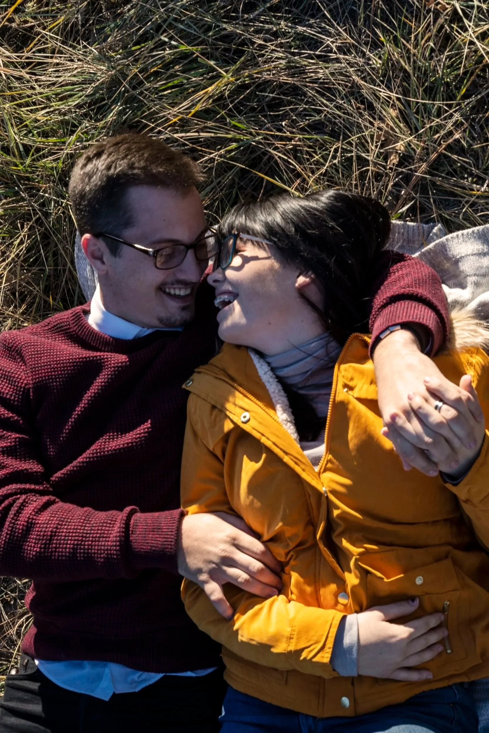 A couple lying outdoors among tall grass, smiling and laughing, embracing each other closely.