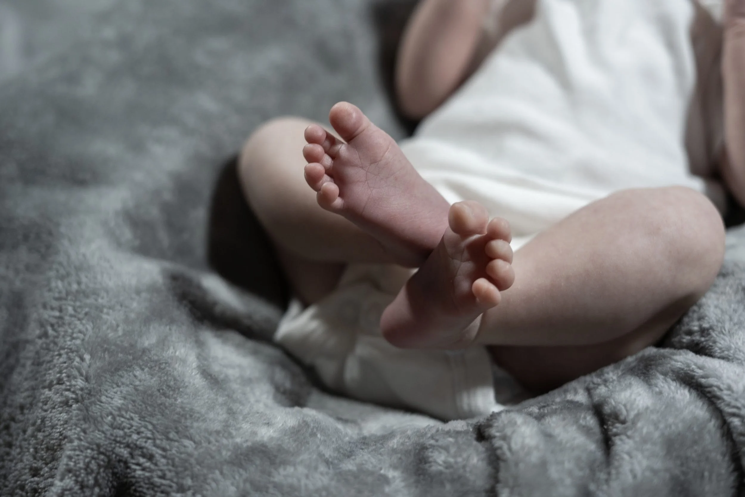 Close-up of a baby's bare feet resting on a soft gray blanket, with part of the baby's legs and a white outfit visible.