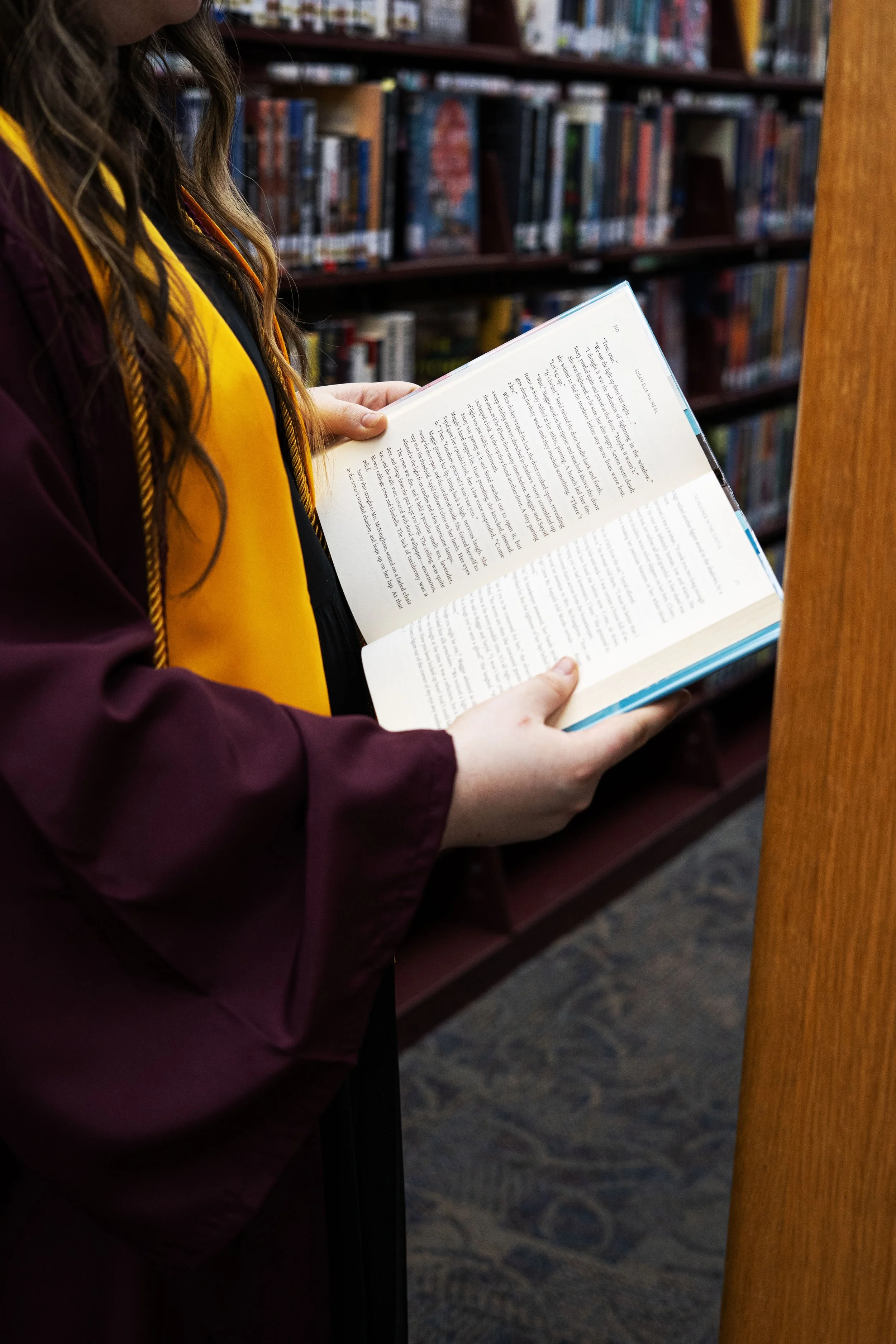 Person in graduation gown reading a book in a library.
