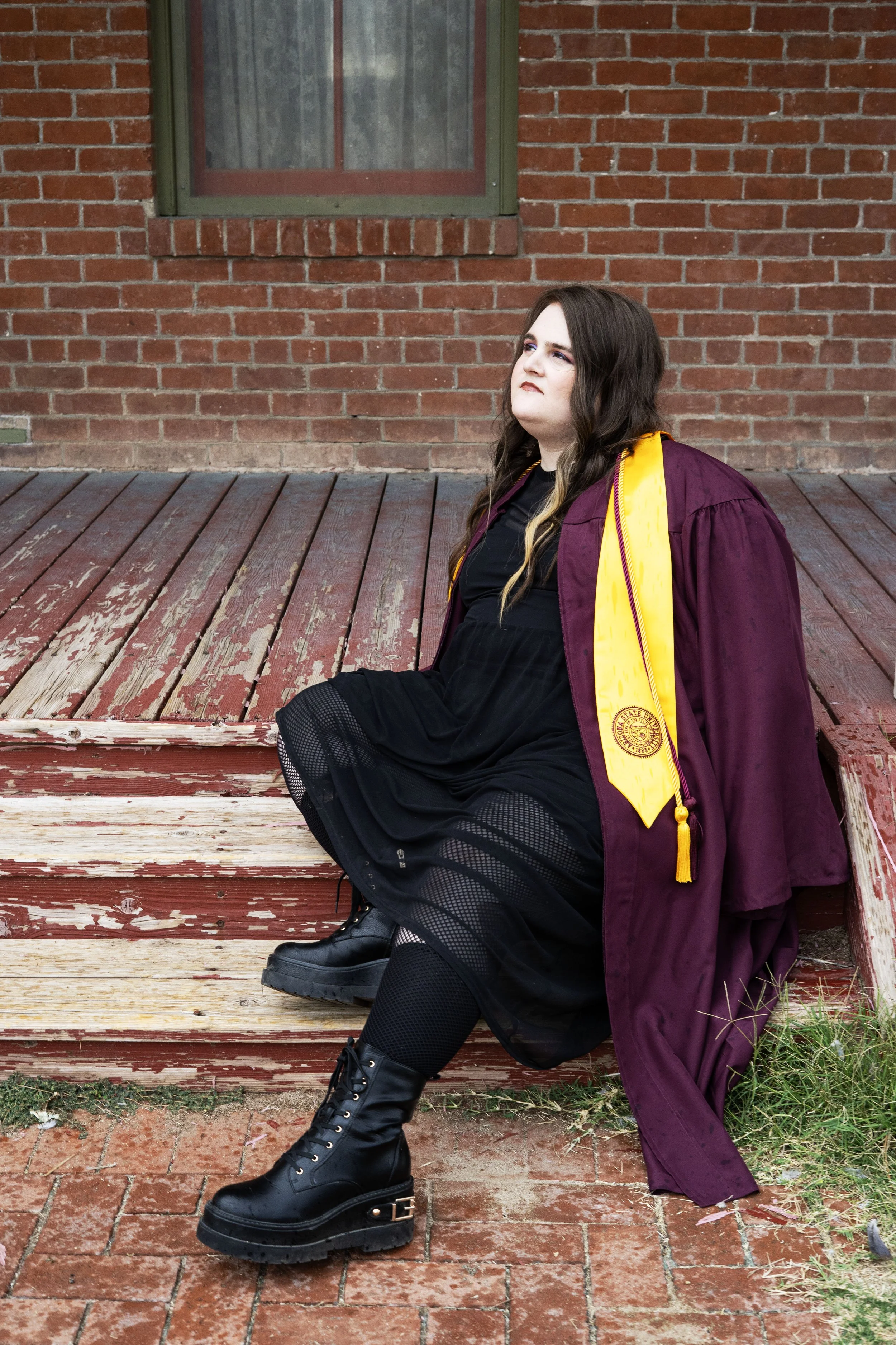 Person with long dark hair sitting on a worn wooden porch, wearing a black dress, black boots, and a maroon and yellow graduation gown and stole, in front of a brick wall with a window.