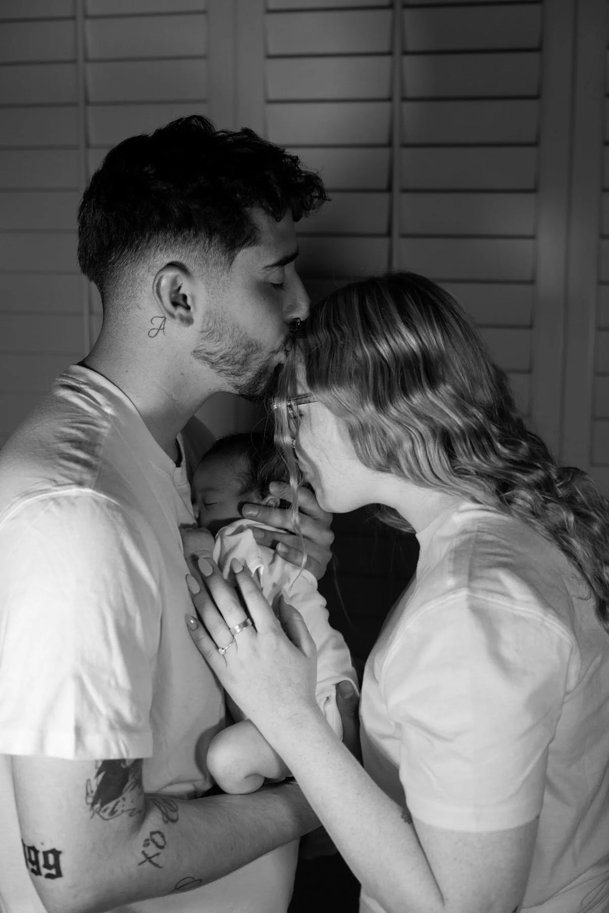 A couple holding a newborn baby, with the father kissing the mother's forehead, in a tender moment.