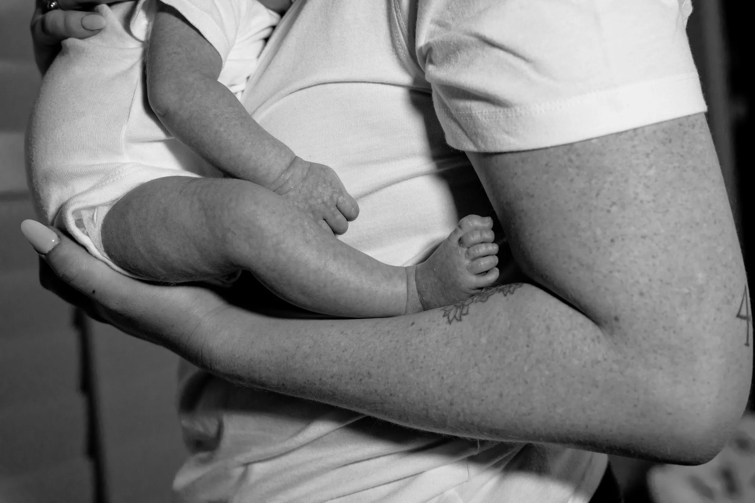 A person holding a newborn baby on their chest, showing baby's tiny hand gripping adult's finger in black and white.