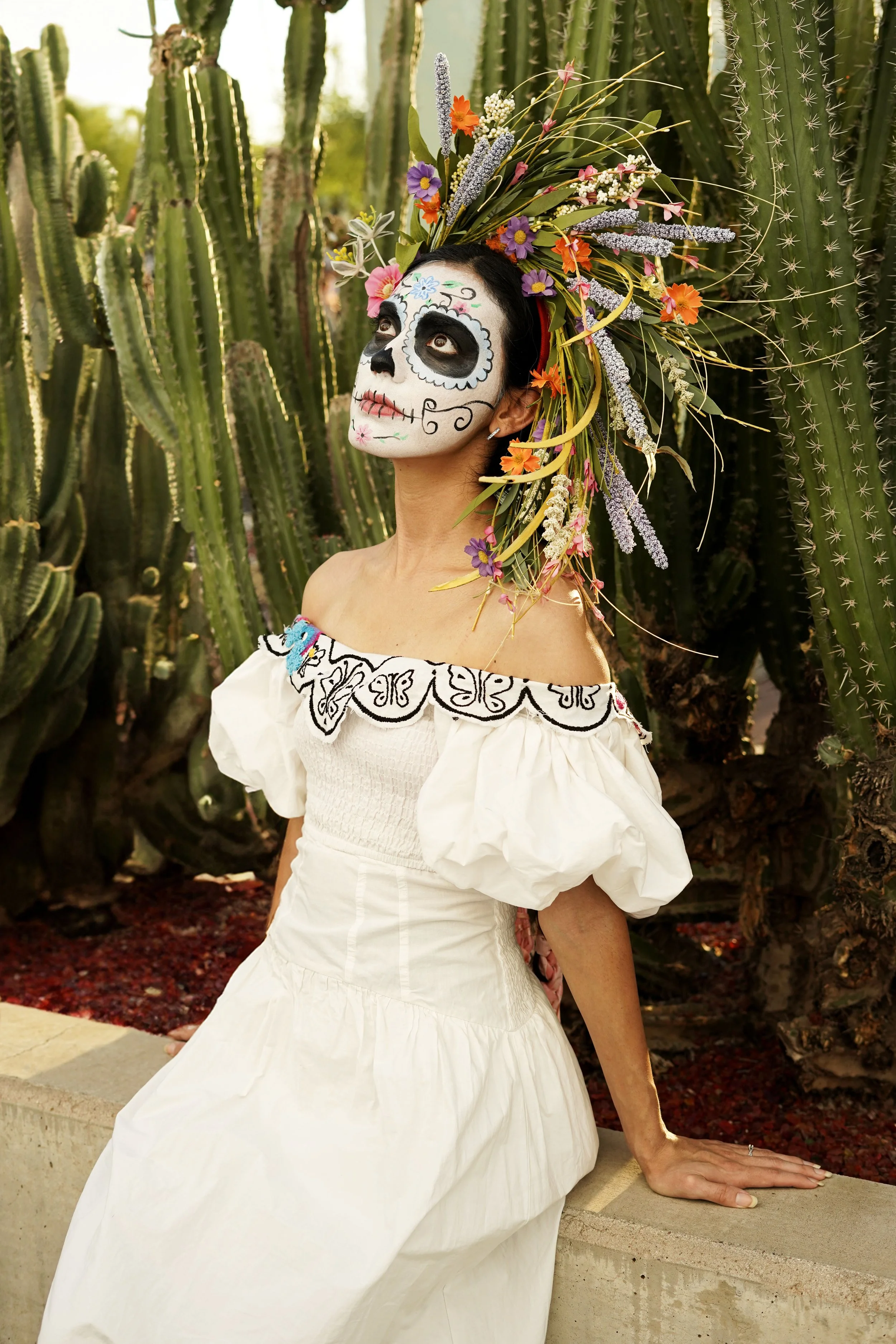 A woman with sugar skull face paint, wearing a white off-shoulder dress with embroidered black details, sits on a low wall among tall cacti, with a large floral headpiece featuring colorful flowers and greenery.
