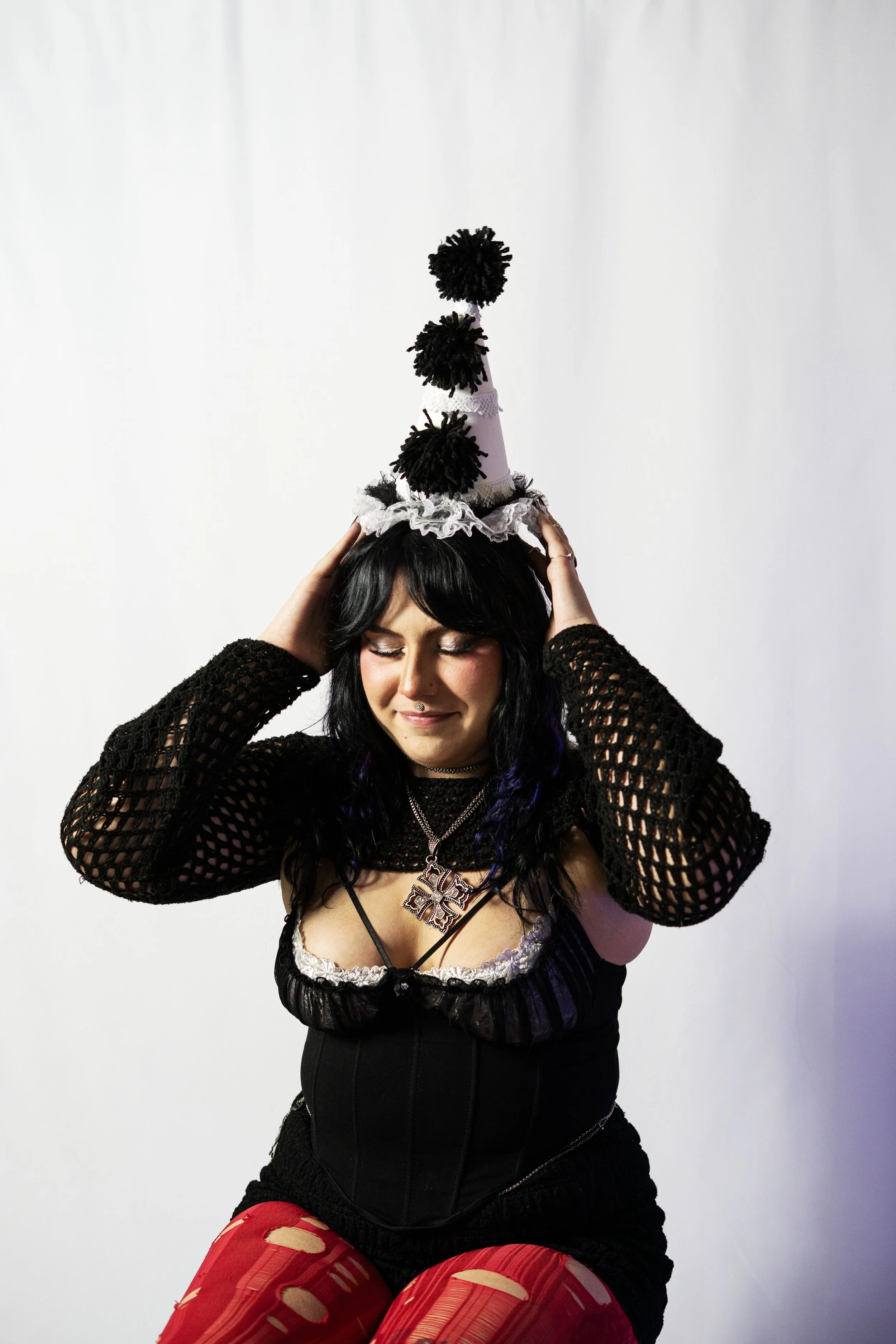 A woman wearing a black lace top, a corset, and red leggings with cut-out designs, is smiling with her eyes closed, holding a black party hat with pom-poms on her head, against a plain white background.