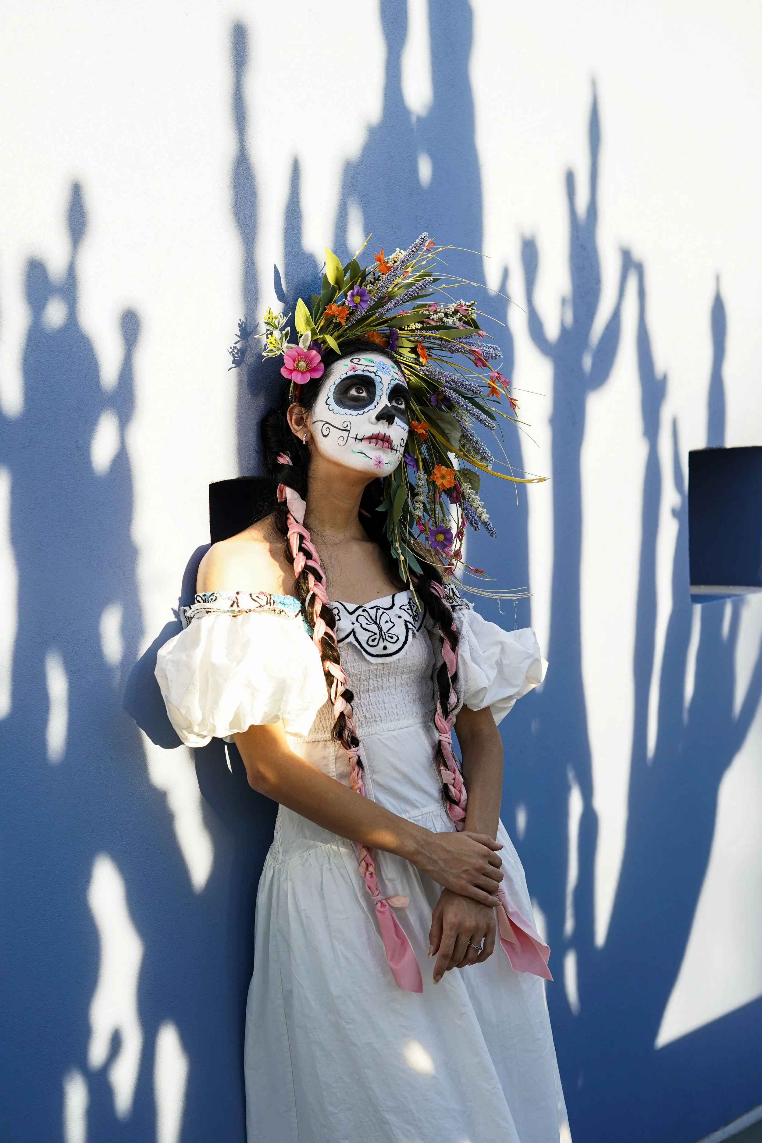 Woman with sugar skull face paint and a floral headdress, wearing an off-shoulder white dress, standing against a blue wall with shadow patterns.