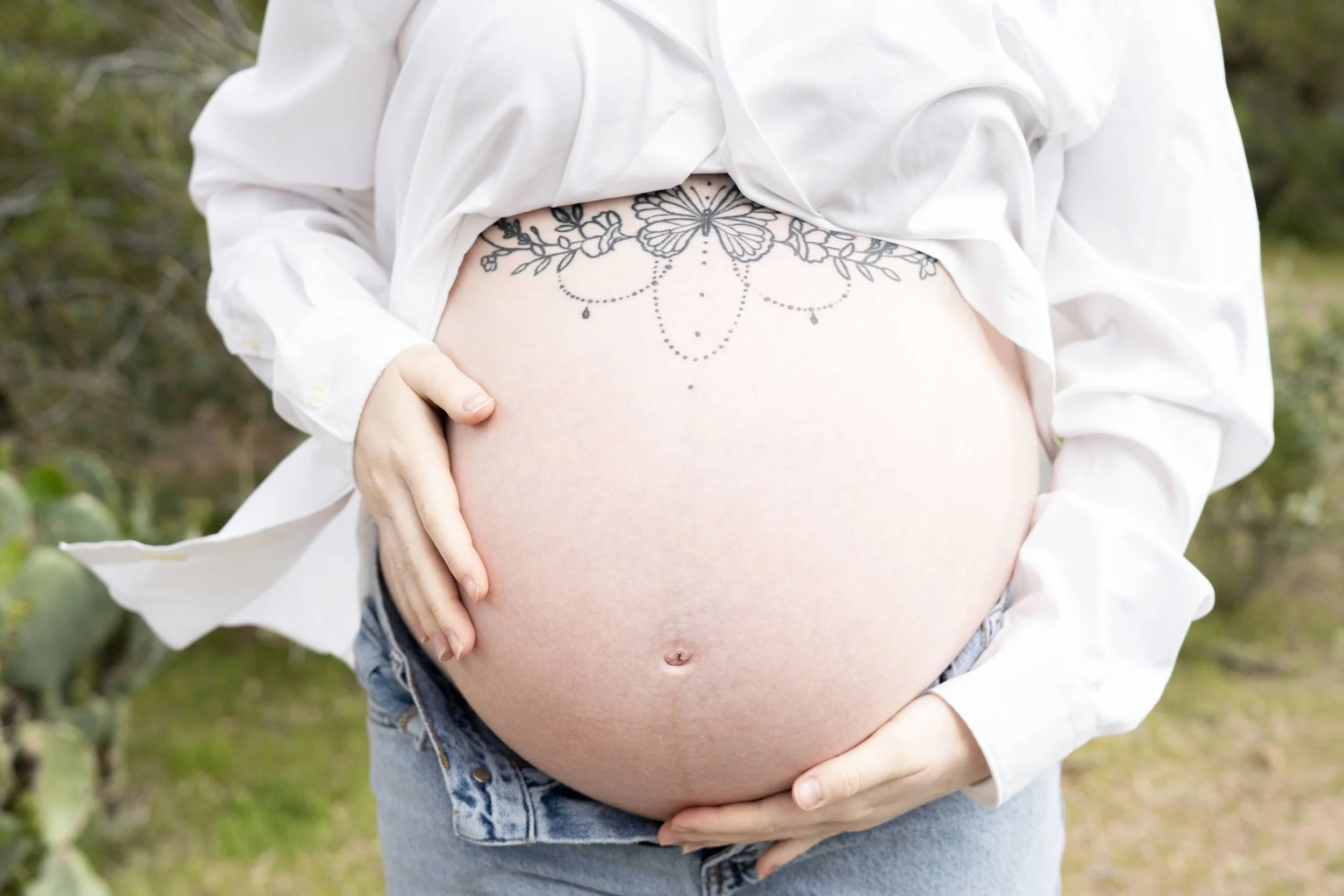 Pregnant woman with a butterfly and floral tattoo on her belly, wearing a white shirt and jeans, outdoors.