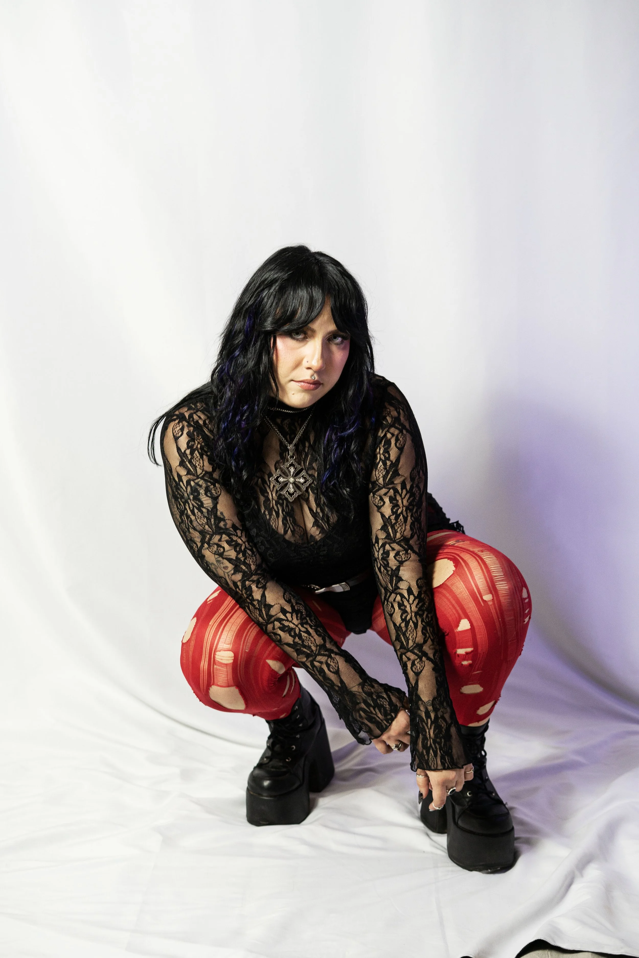A woman with long black hair with purple highlights, wearing a black lace top, red ripped pants, and black platform boots, squatting on a white backdrop.