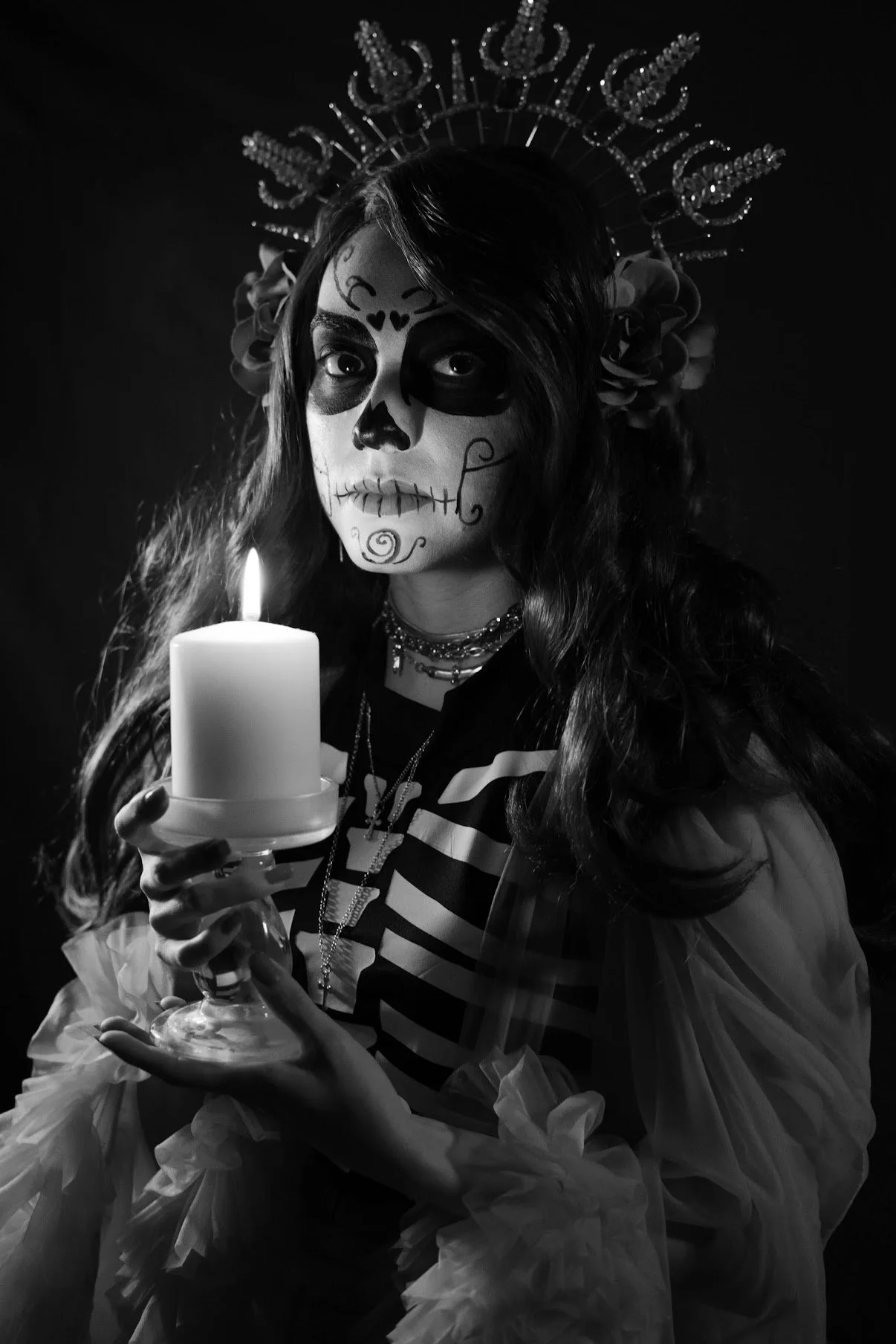 A person with face makeup resembling a sugar skull, holding a lit candle, wearing a crown, flowers, and layered necklaces, dressed in a striped shirt and tulle sleeves, in black and white.