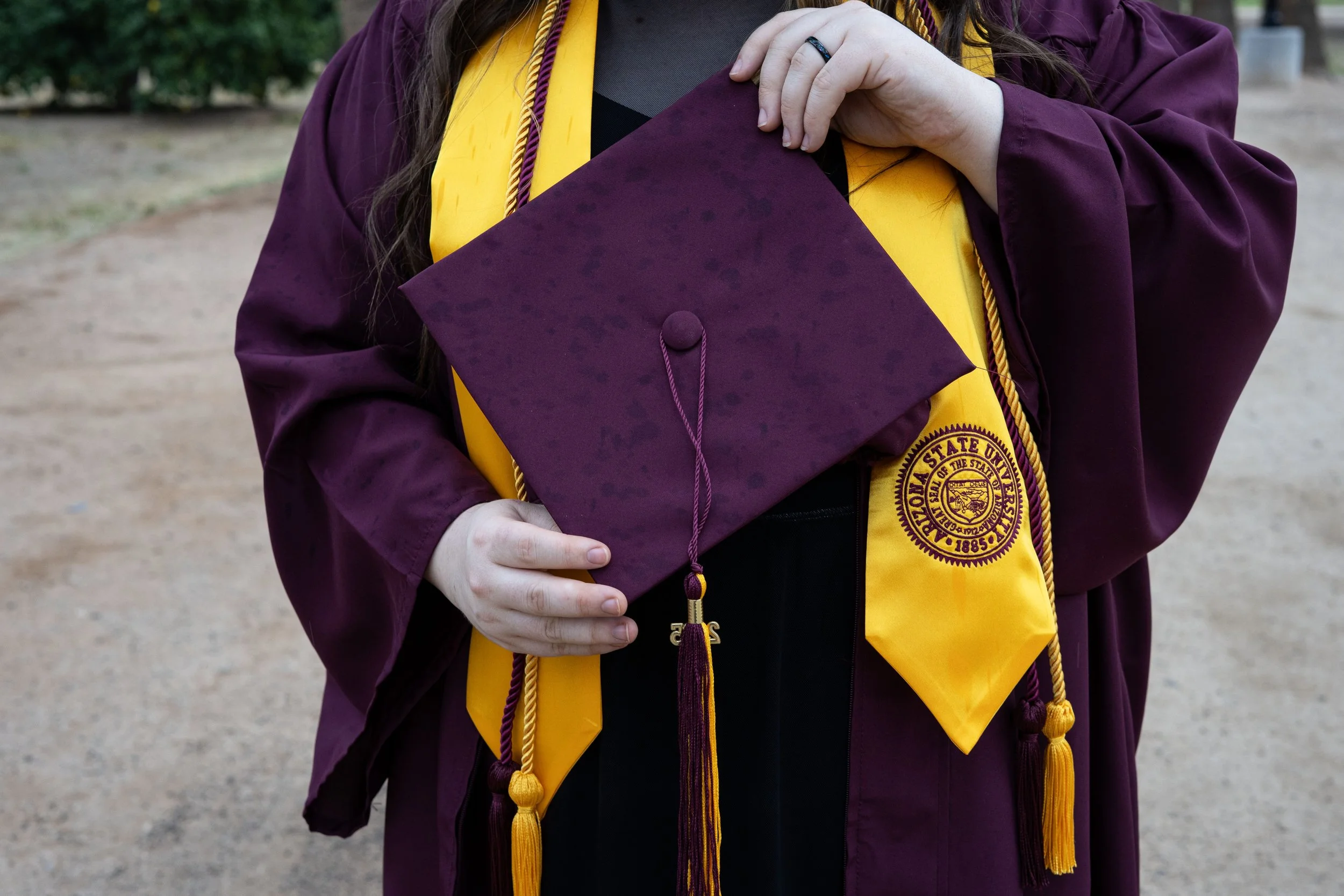 A person in a maroon graduation gown holding a maroon cap, with a yellow stole and tassels, standing outdoors.