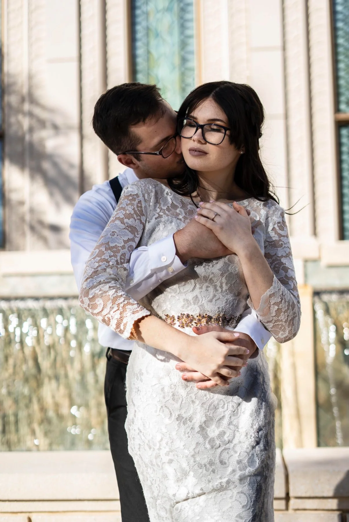A man in a white shirt and glasses embraces a woman in a white lace dress and glasses outdoors, with her hands over her chest and a serious expression.