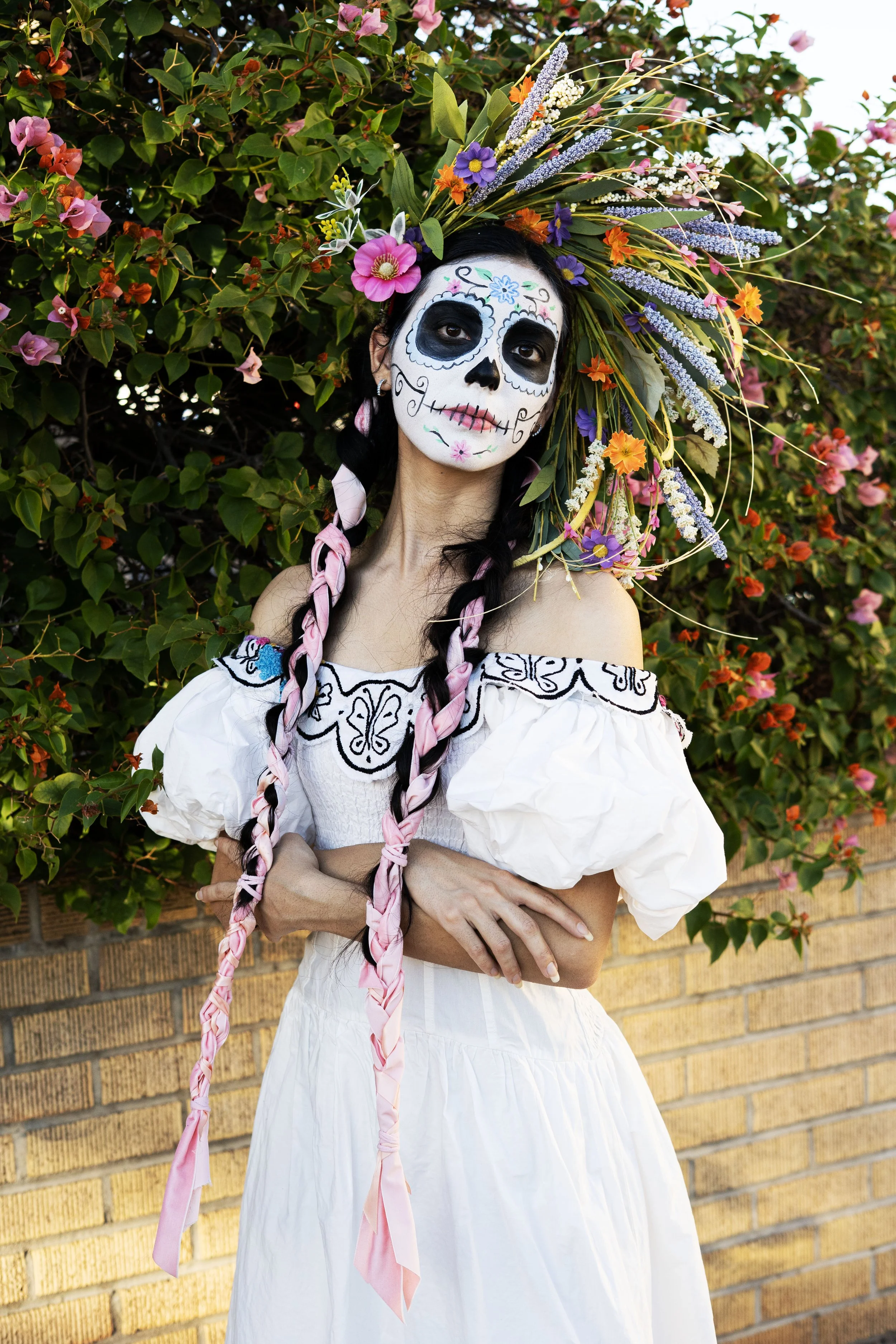 A woman dressed in traditional Mexican attire with sugar skull face paint, wearing a floral headdress, standing outdoors in front of a brick wall and green bushes with colorful flowers.