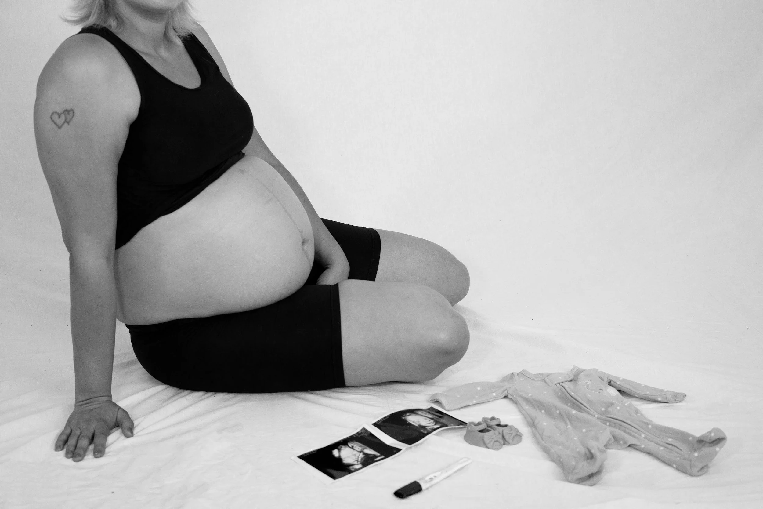 Pregnant woman sitting on the floor with ultrasound images, a baby outfit, socks, and a marker nearby, black and white photo.
