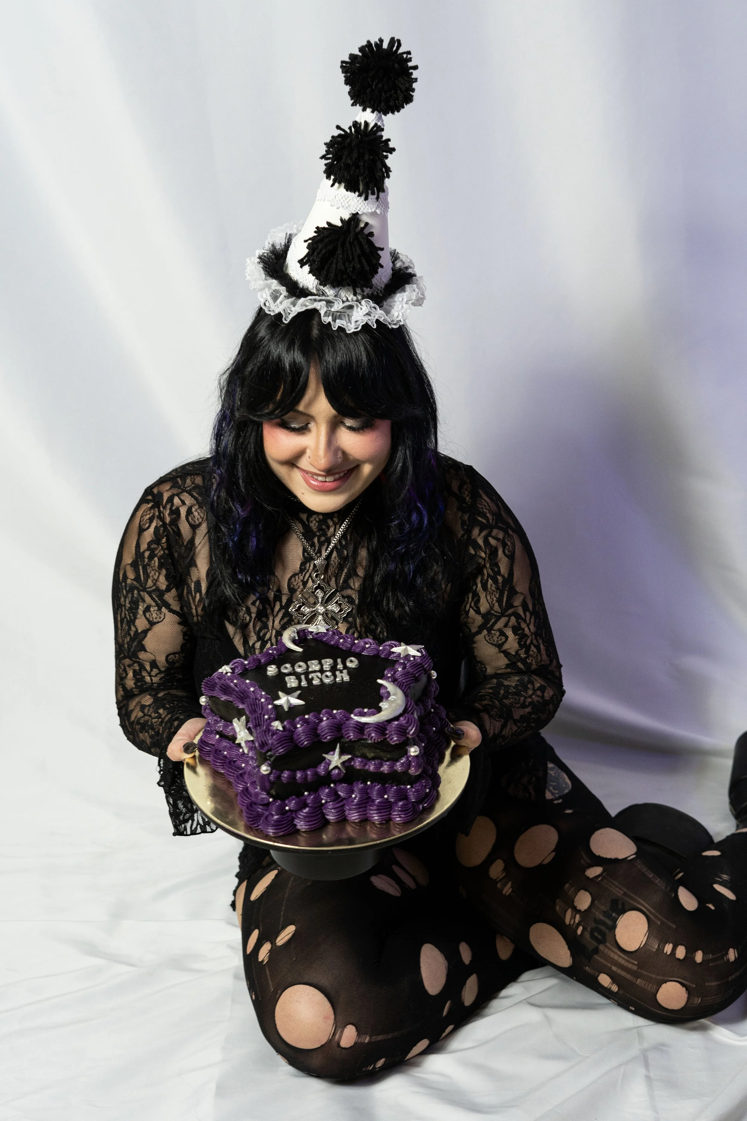 Woman with black hair wearing a black lace top, fishnet tights, and a ruffled party hat, holding a purple birthday cake decorated with stars and moons, smiling downward.