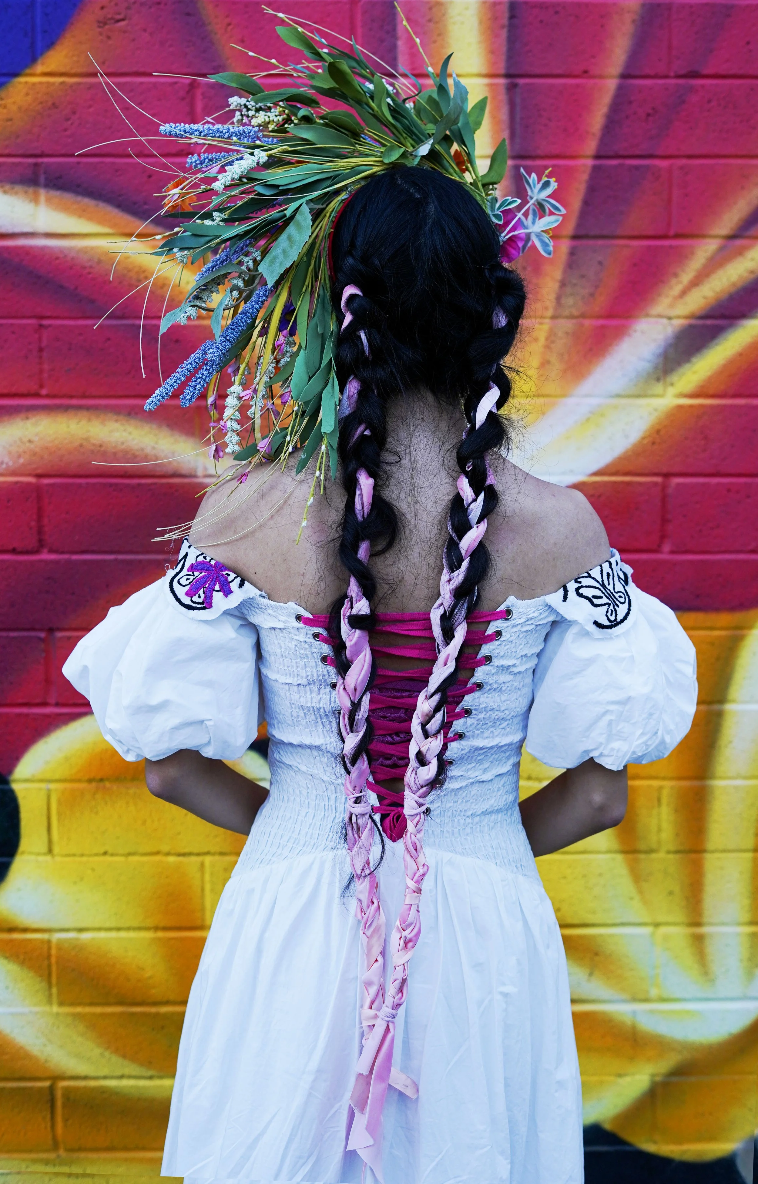 Back view of a woman in a white off-shoulder dress with embroidered butterfly details, wearing braided hair with pink ribbons, standing against a colorful graffiti wall with a floral headdress made of leaves and flowers.