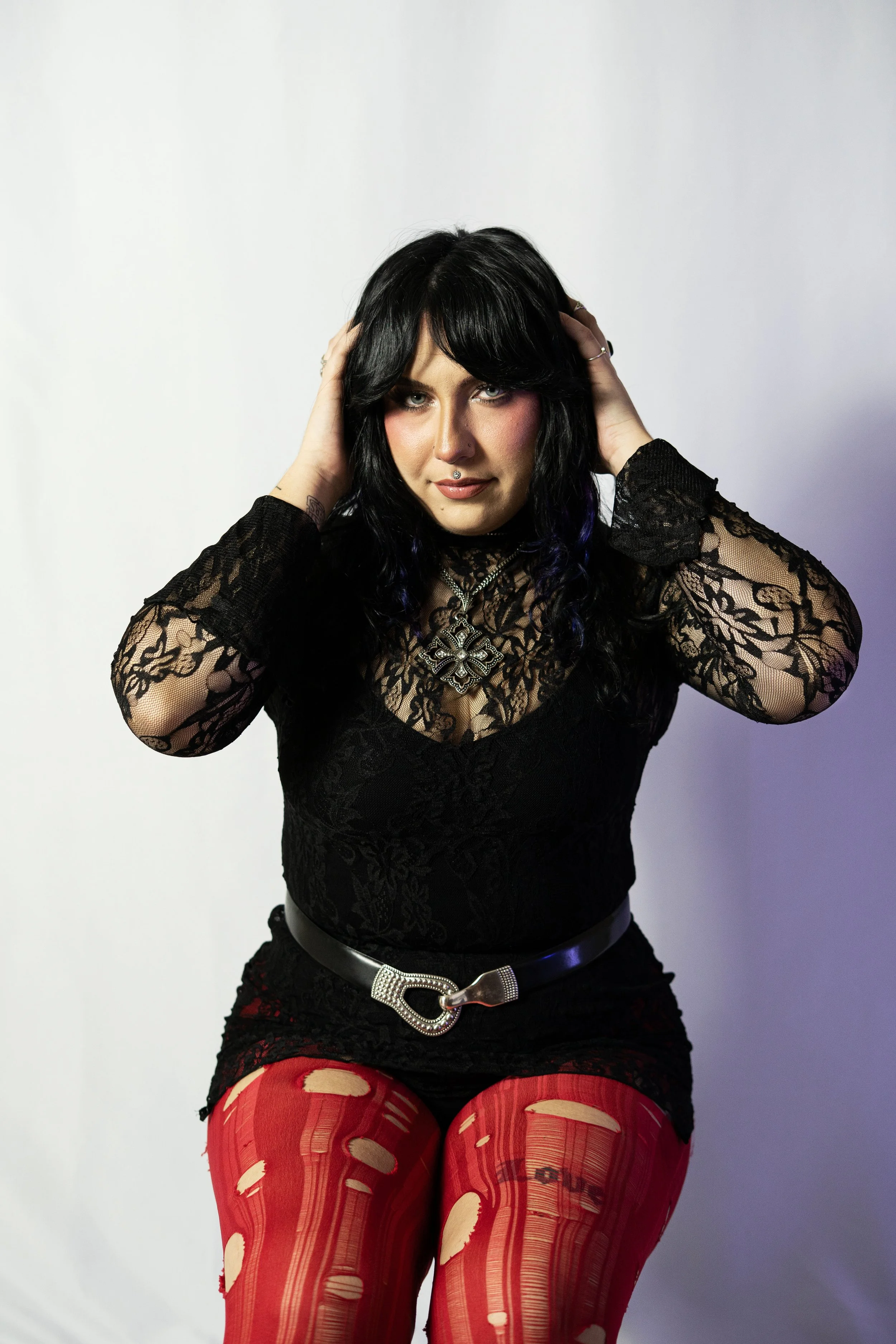 A woman with black hair, with purple streaks, in a black lace top and red ripped tights, sitting against a white background, posing with hands in her hair.