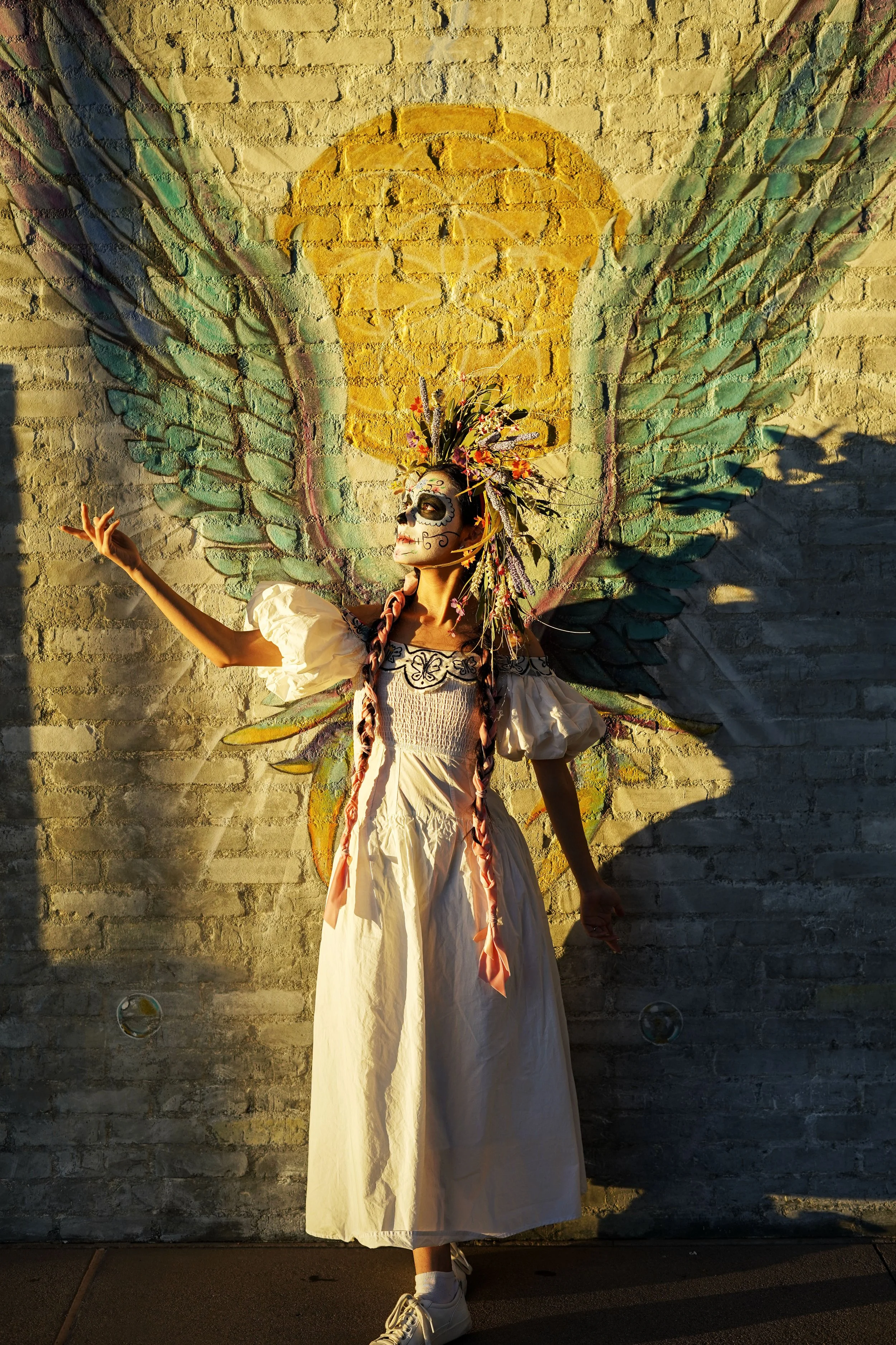 A person dressed in a white gown with colorful accessories and face paint, standing in front of a wall painting of a yellow sun with green wings. The person is wearing a headdress adorned with flowers and decorated with face paint resembling a sugar skull, and their pose resembles a ballet stance.