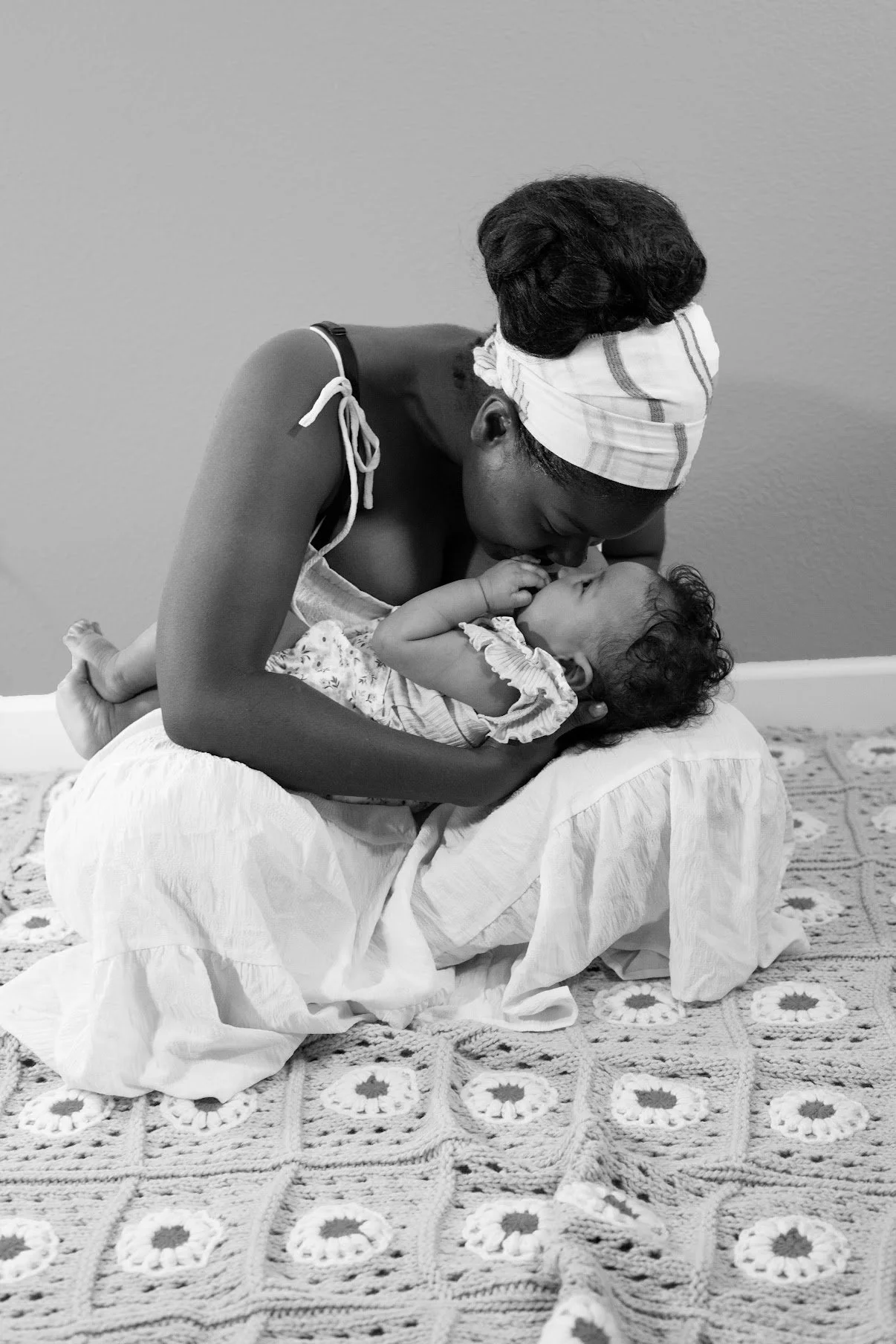 A woman with dark hair and a headscarf bends over a small child lying on a blanket on the floor, touching foreheads as if about to kiss.