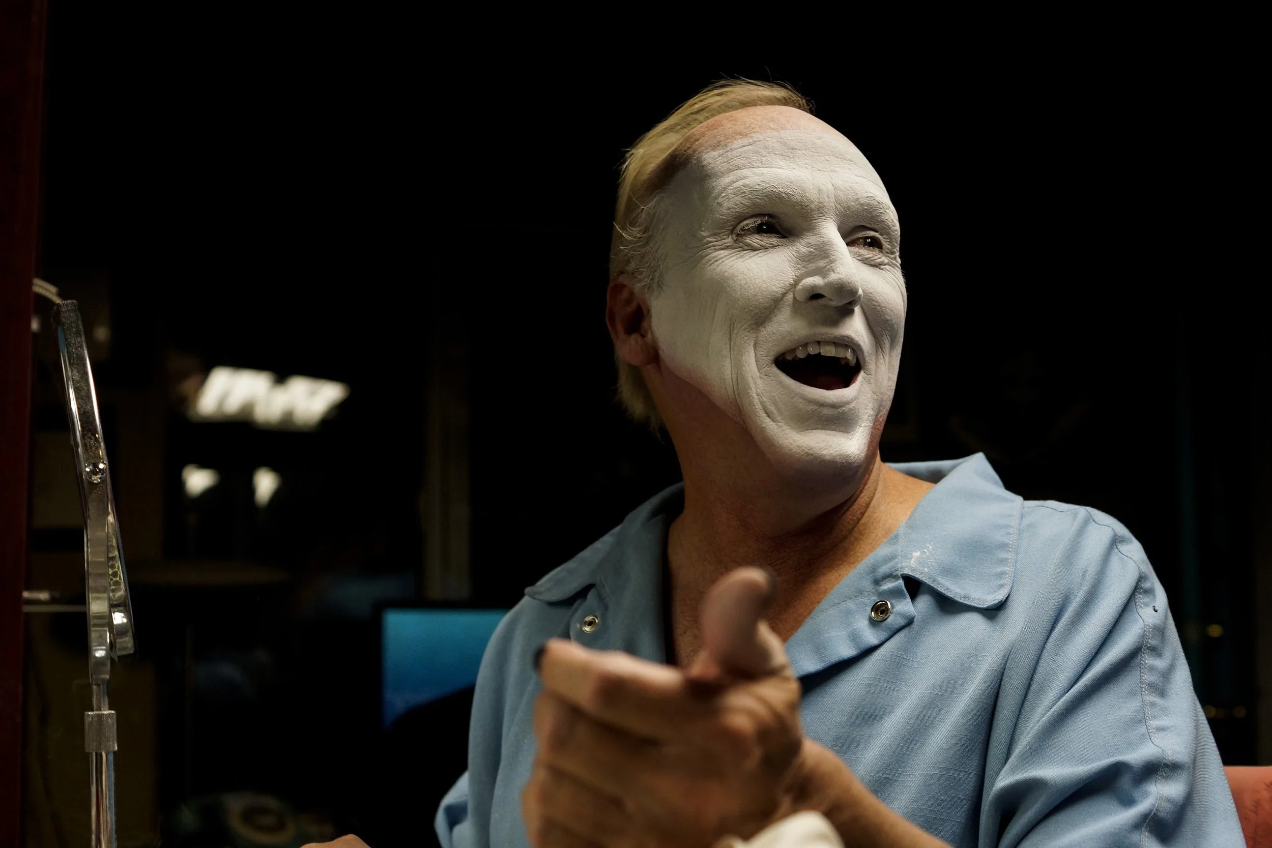 A man with white face makeup, blonde hair, blue shirt, mid-conversation, pointing with his right hand in a dark room.