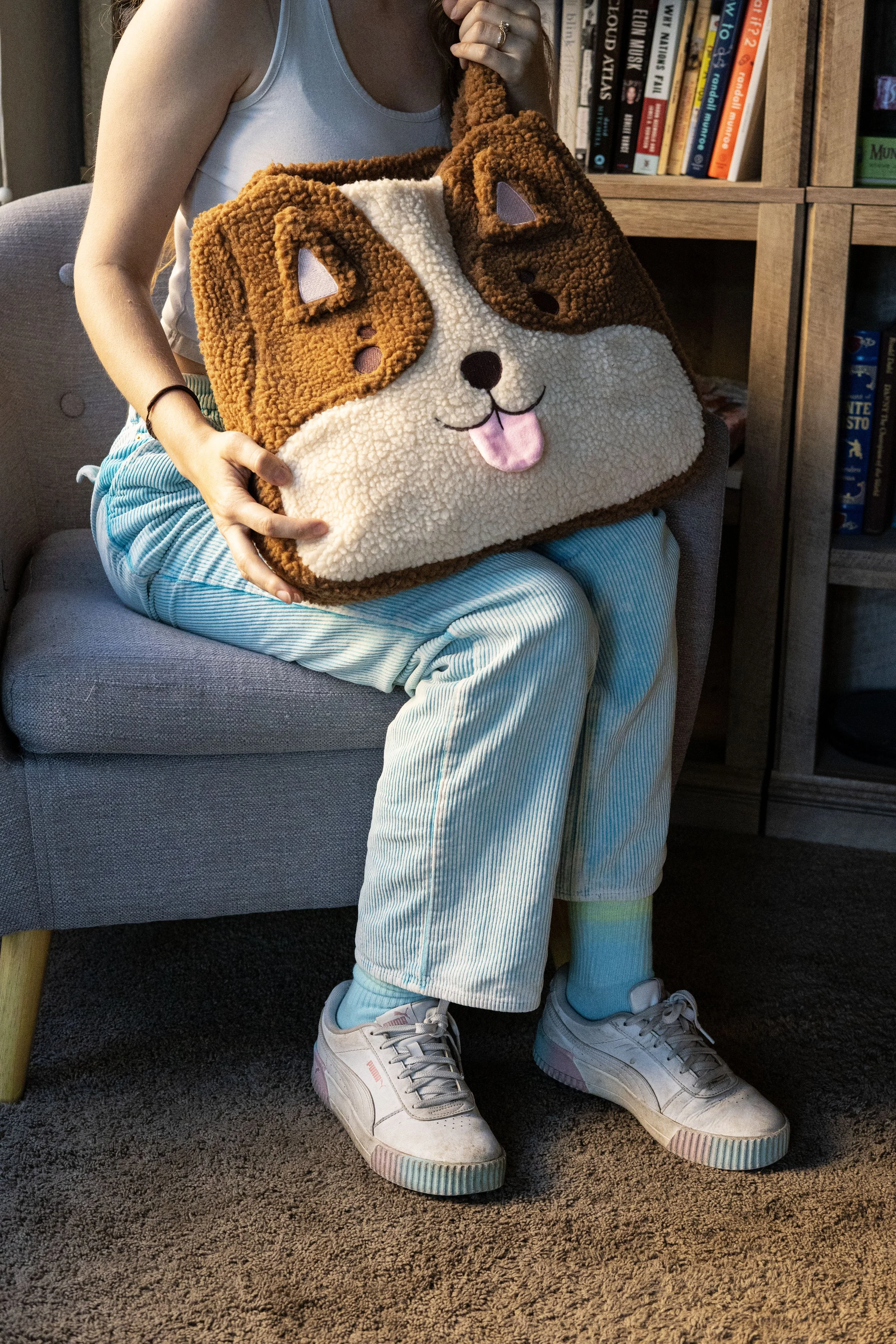 Person sitting on a gray armchair holding a large plush bag that looks like a dog's face, with a background of a bookshelf filled with books.